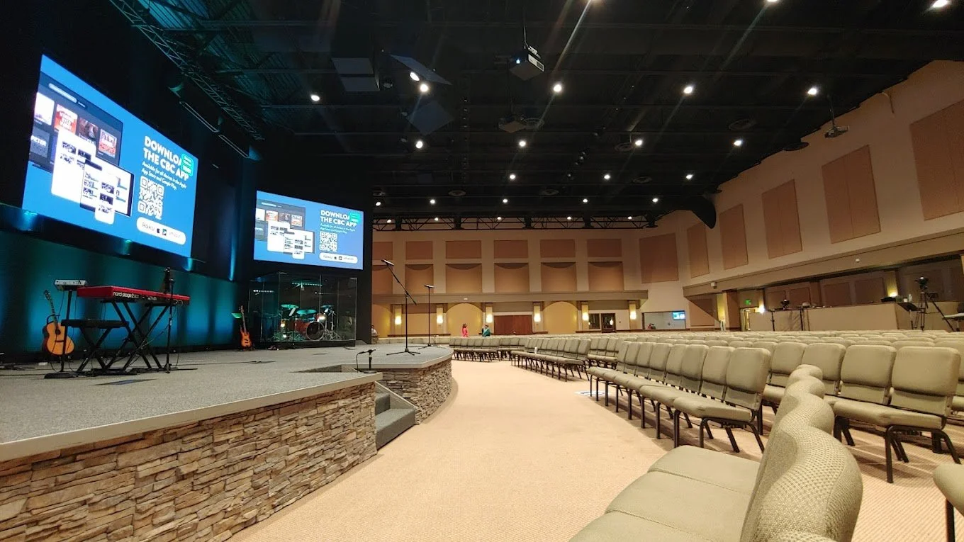 Empty auditorium with a stage, musical instruments, large screens displaying an app download, and rows of beige chairs.