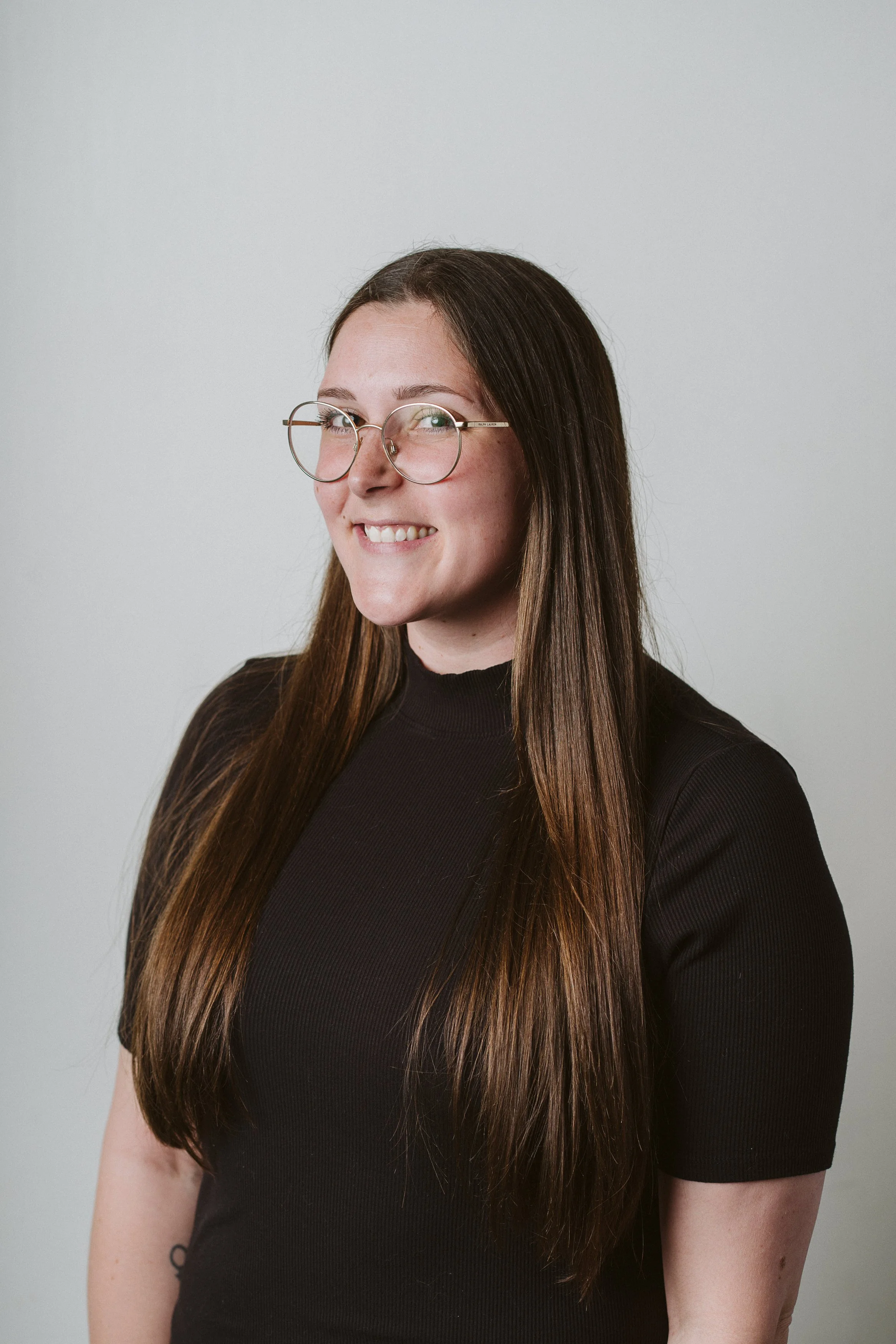 A woman with long brown hair wearing glasses and a grey sweater, smiling, against a plain light grey background.