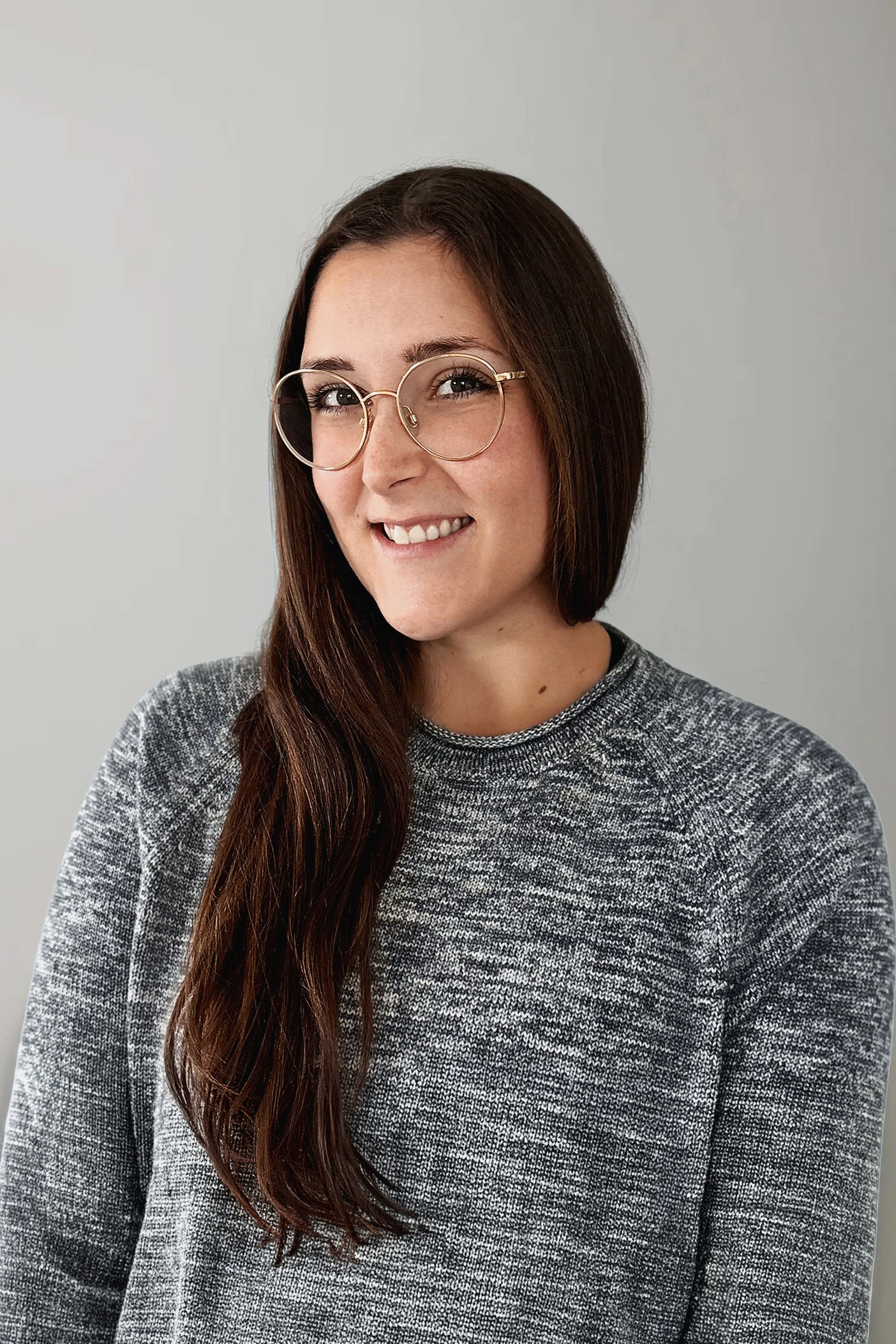 A young woman with long brown hair, wearing round glasses and a gray patterned sweater, smiling and looking at the camera against a plain gray background.