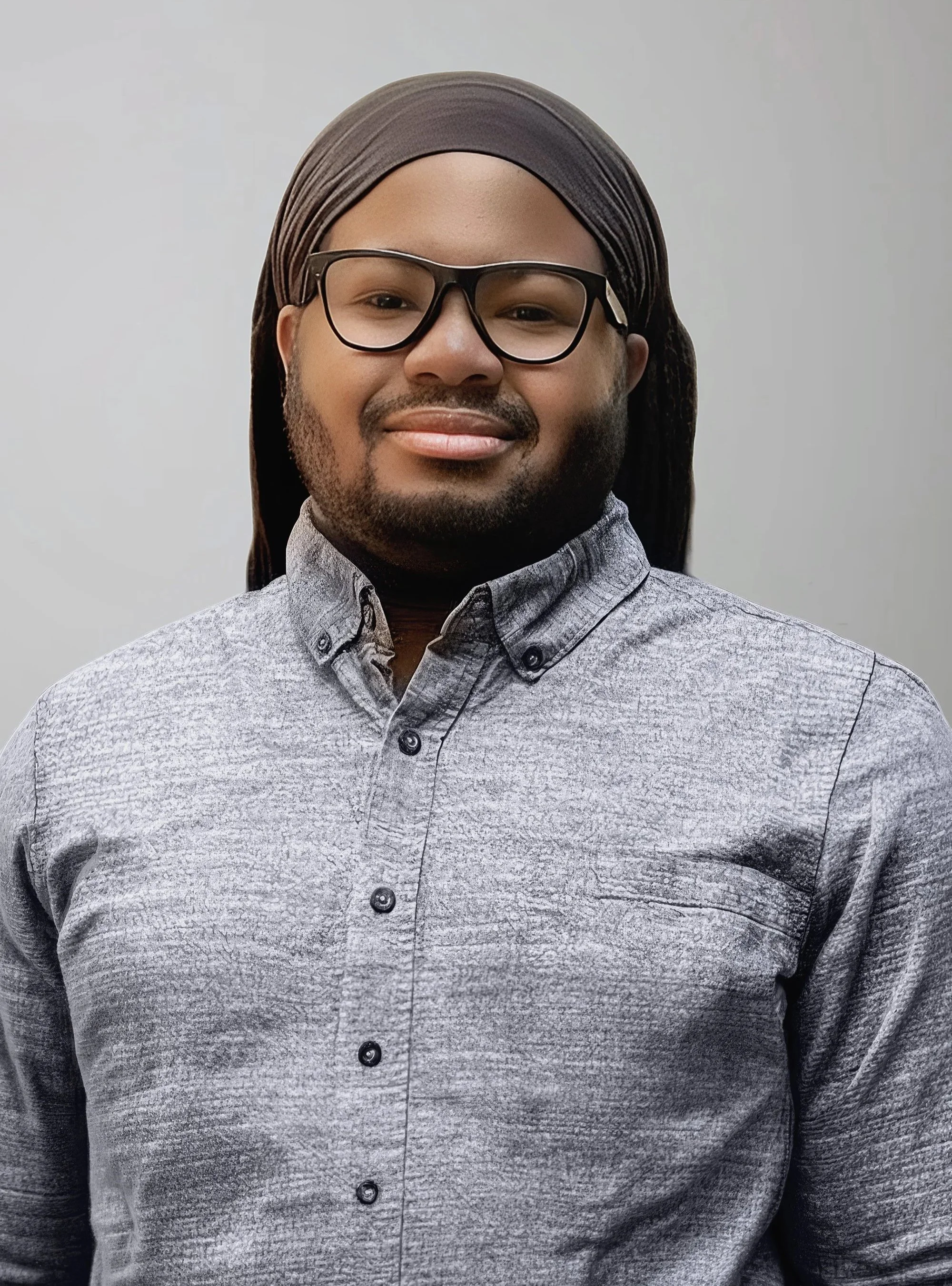 A man wearing glasses, a brown head covering, and a gray patterned button-up shirt is smiling and looking at the camera against a plain light-colored background.