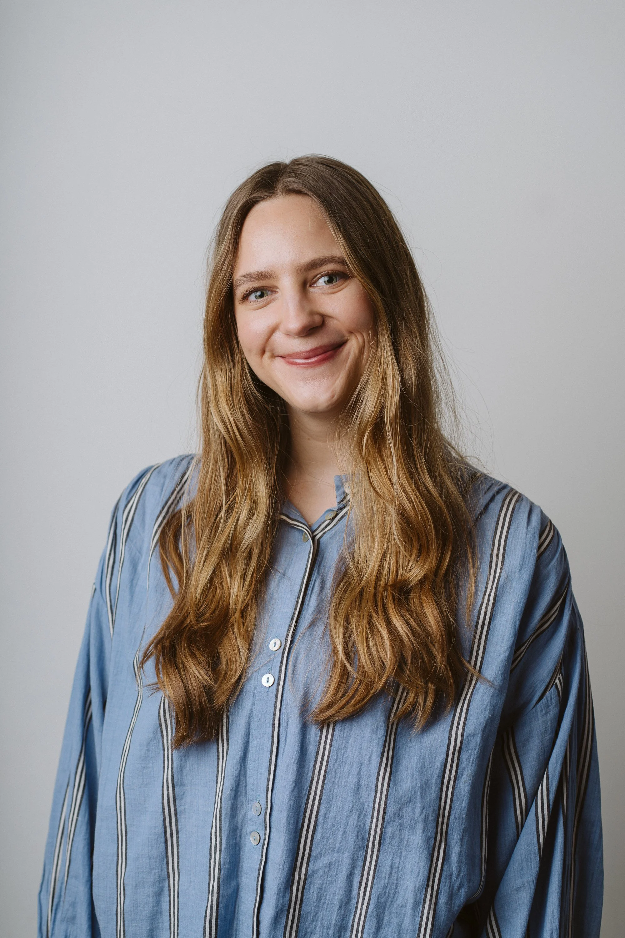 A woman with long, wavy blonde hair wearing a light blue button-up shirt with dark vertical stripes, smiling softly against a plain light gray background.