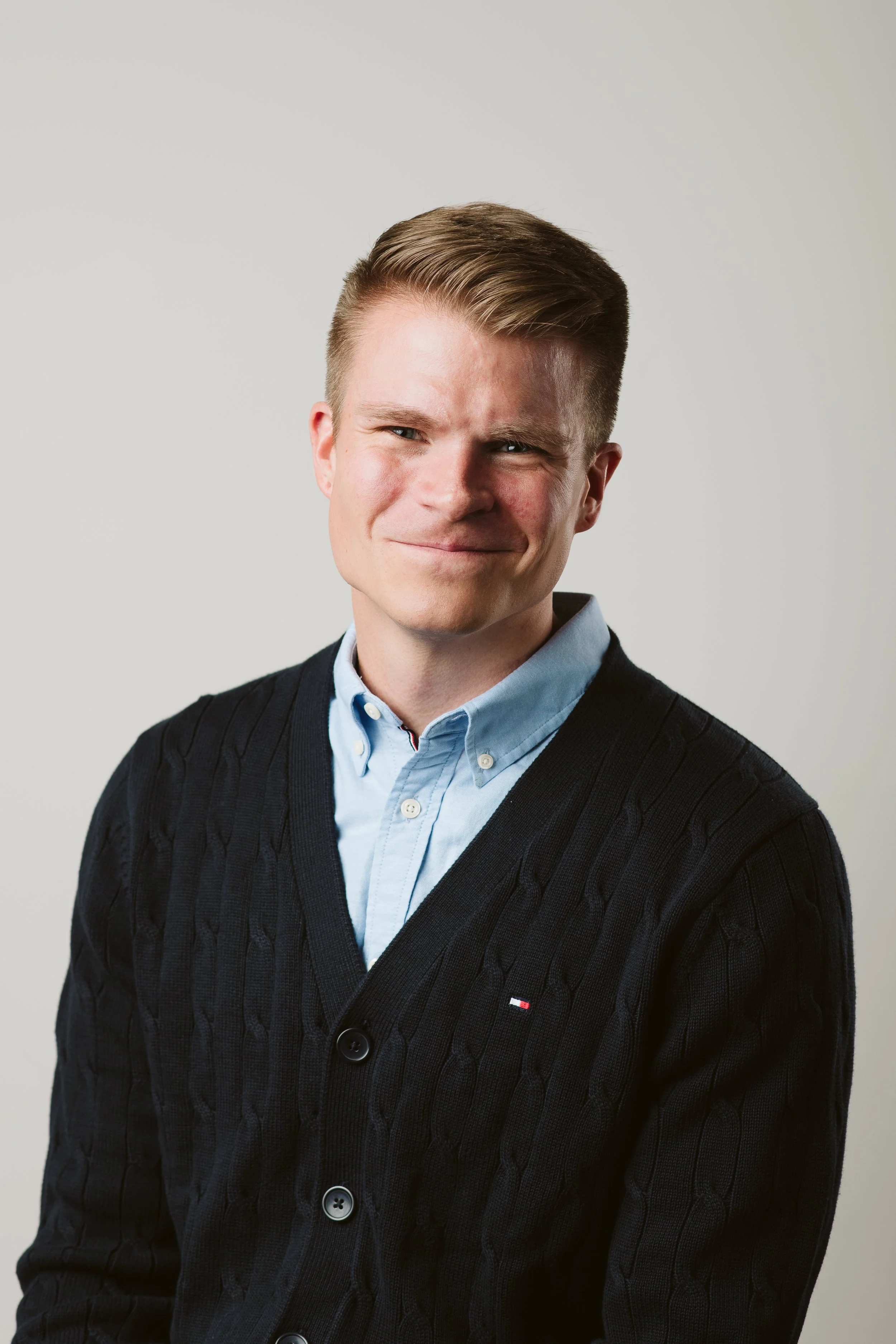 Portrait of a young man with short, styled hair, wearing a light blue collared shirt and a black knit cardigan with small buttons, smiling subtly against a plain light gray background.