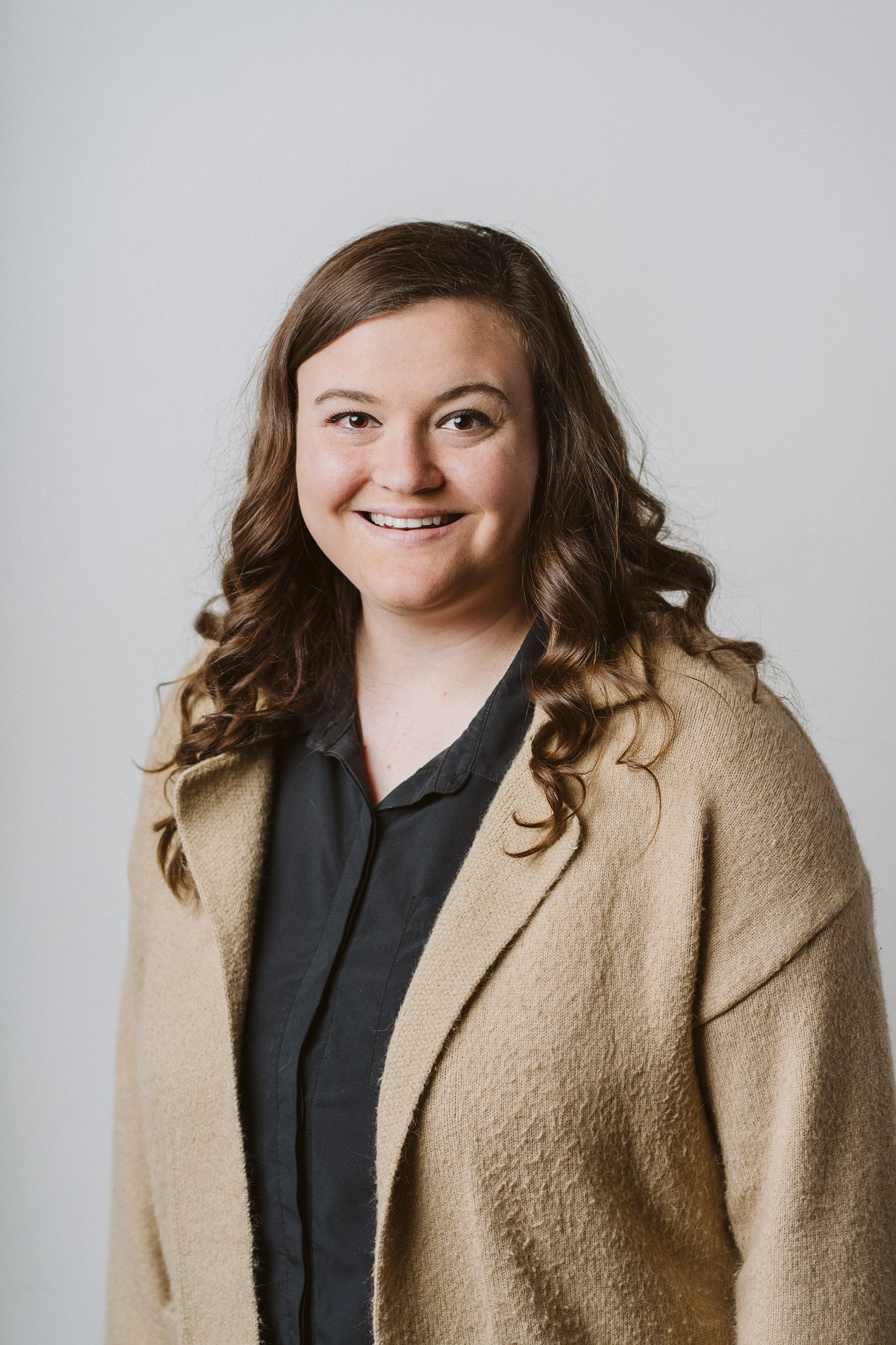 Headshot of a woman with brown curly hair, smiling, wearing a black shirt and a beige coat, against a plain light gray background.