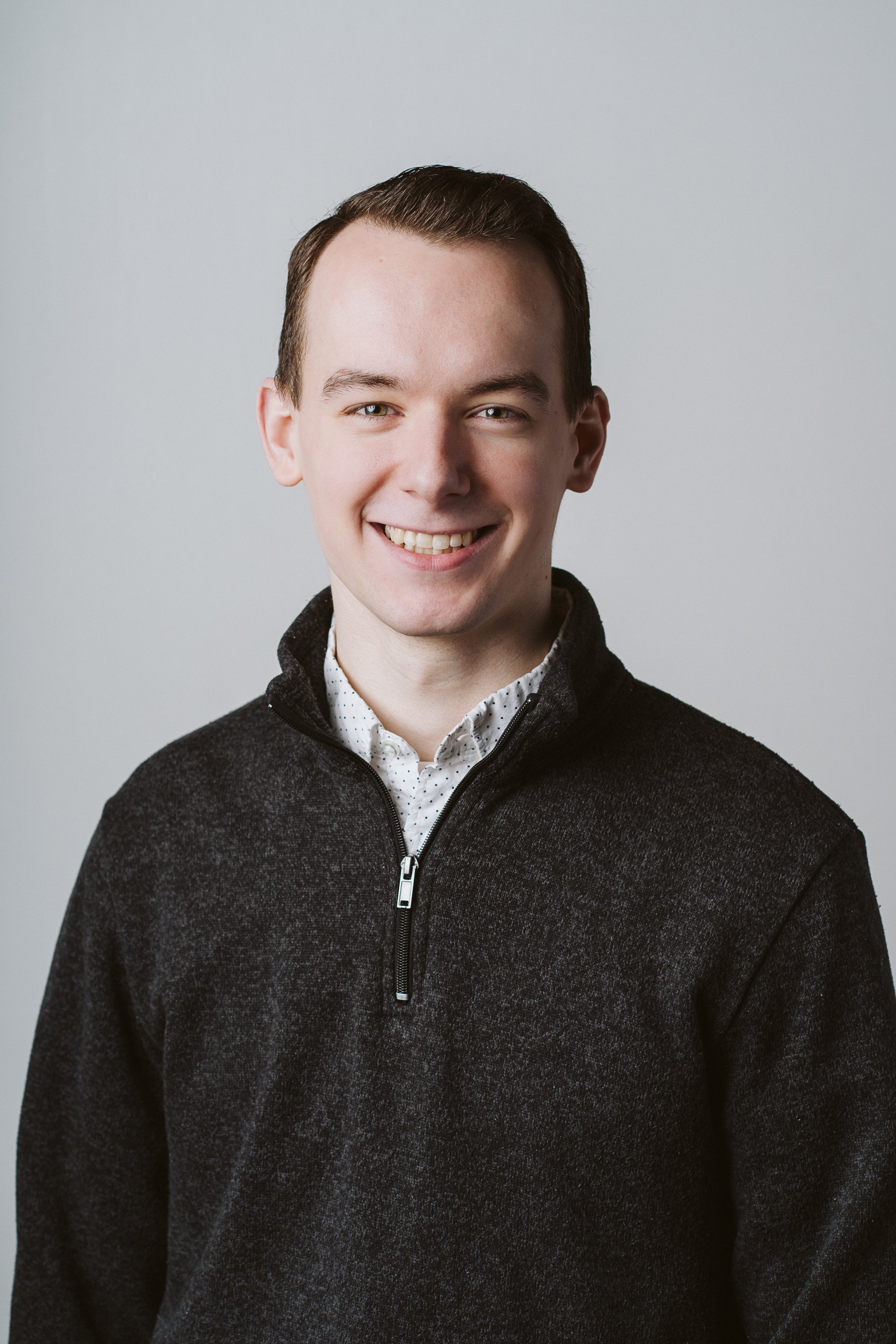 Smiling young man with short brown hair, wearing a white shirt with small black dots and a black zip-up sweater, against a plain light gray background.