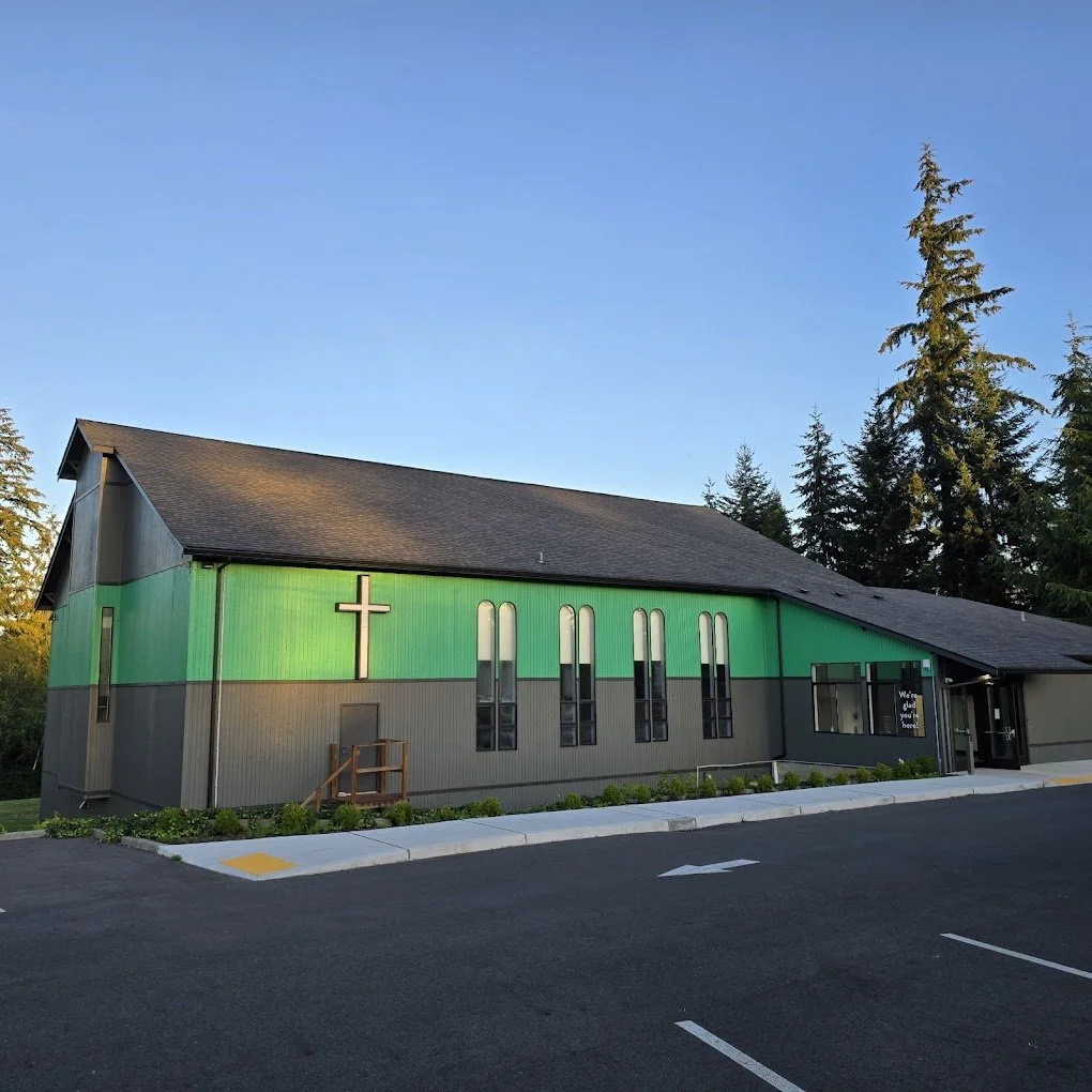 A church building with a green facade, a wooden cross, tall narrow windows, and trees in the background under a blue sky.