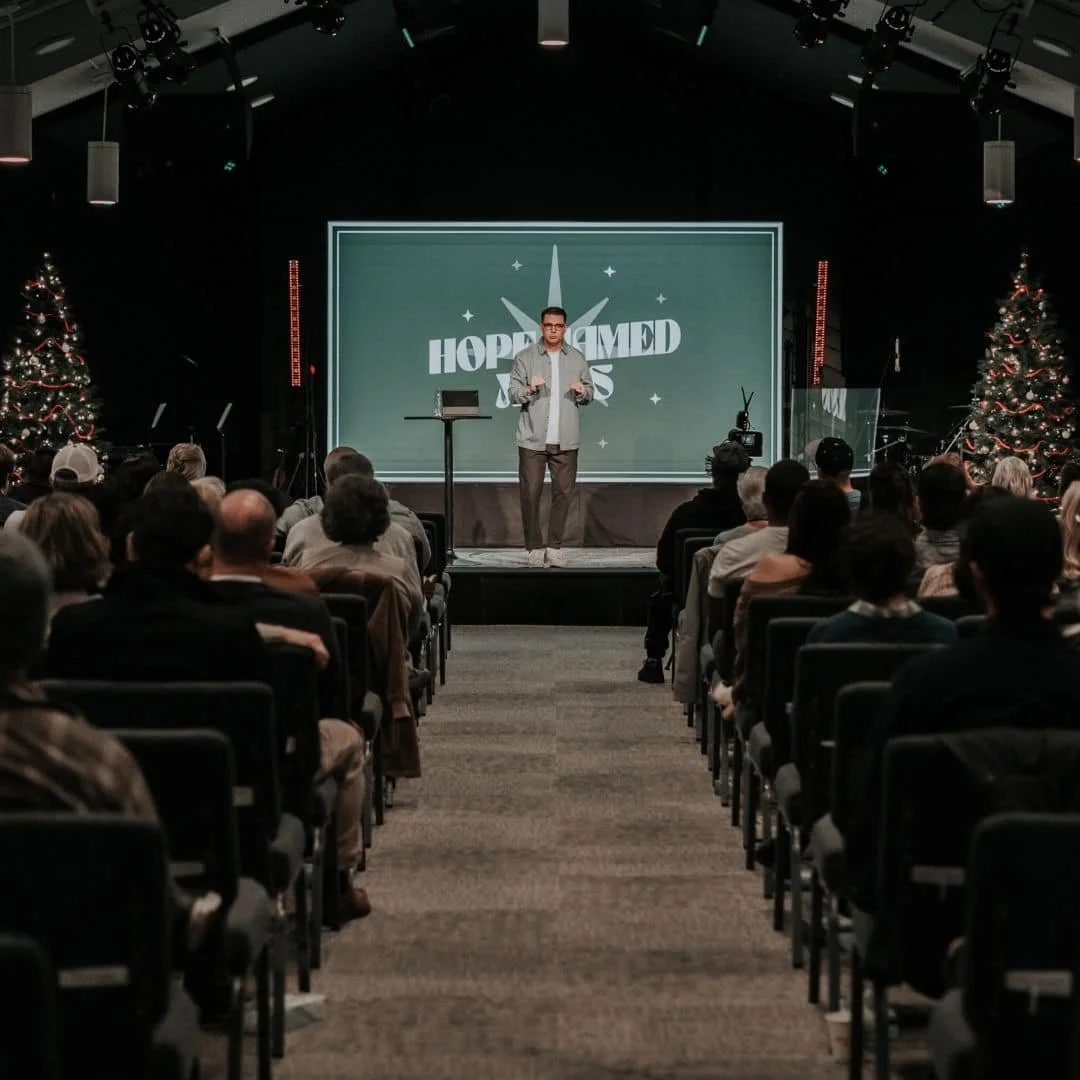 A man speaking on stage at a religious event called 'HOPEDMED JESUS' in a dark auditorium decorated with Christmas trees, with an audience seated facing the stage.