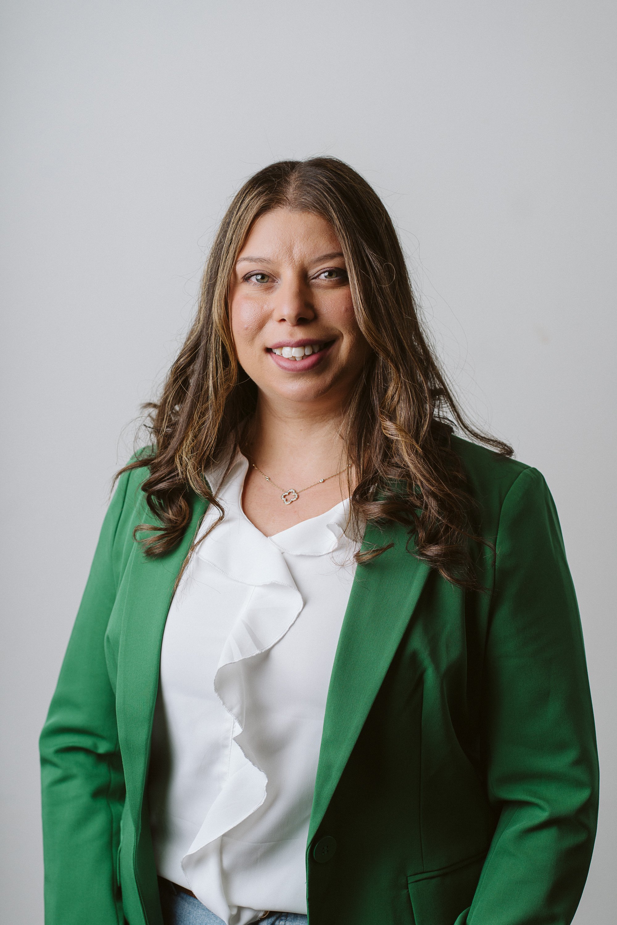 A woman with long wavy brown hair and blue eyes smiling outdoors, wearing a light gray blazer and white blouse with a gold necklace.