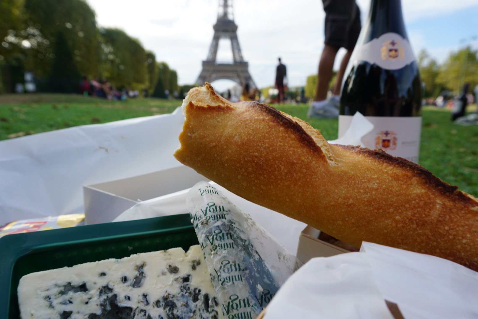 Parisian travel-inspired oil painting featuring a picnic of baguette, cheeses, butter, pastries, and wine beneath the Eiffel Tower. Food art by Courtney Caroline
