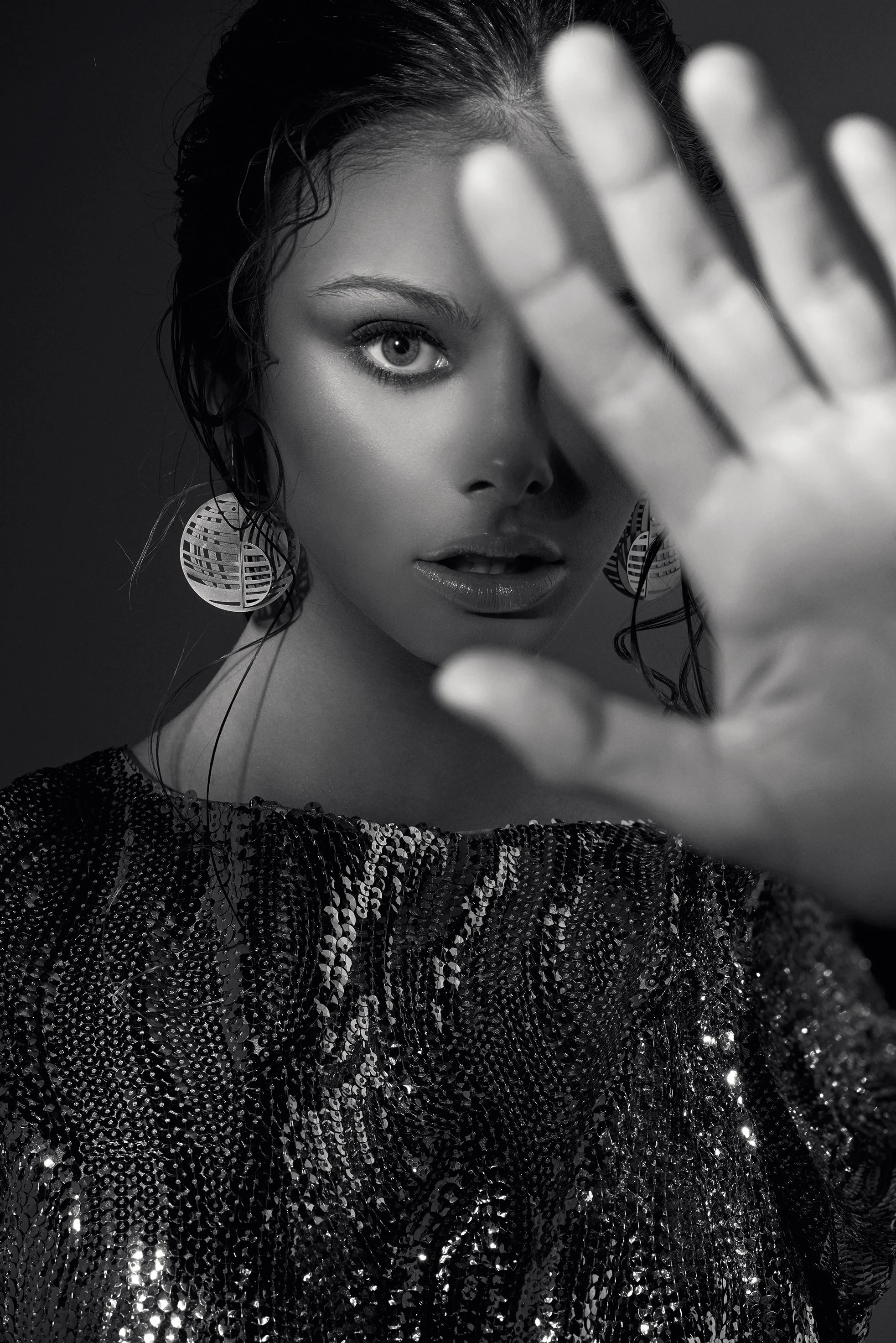 Black and white portrait of a woman with wet hair, wearing large circular earrings and a sequin top, holding her hand up towards the camera, partially covering her face.