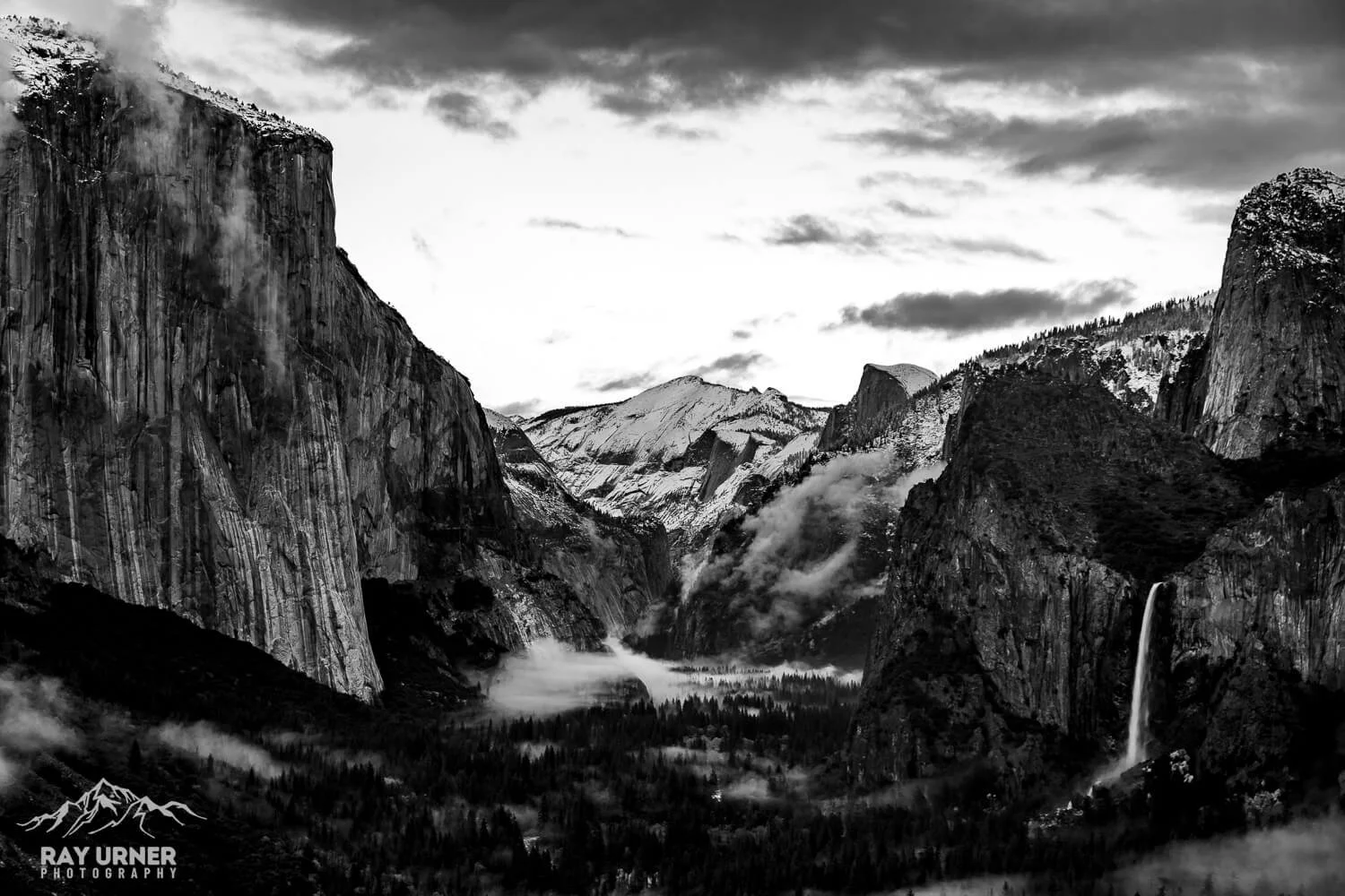 Black and white photograph of Yosemite Valley, featuring tall granite cliffs, snow-capped mountains, a waterfall, and forested valley.