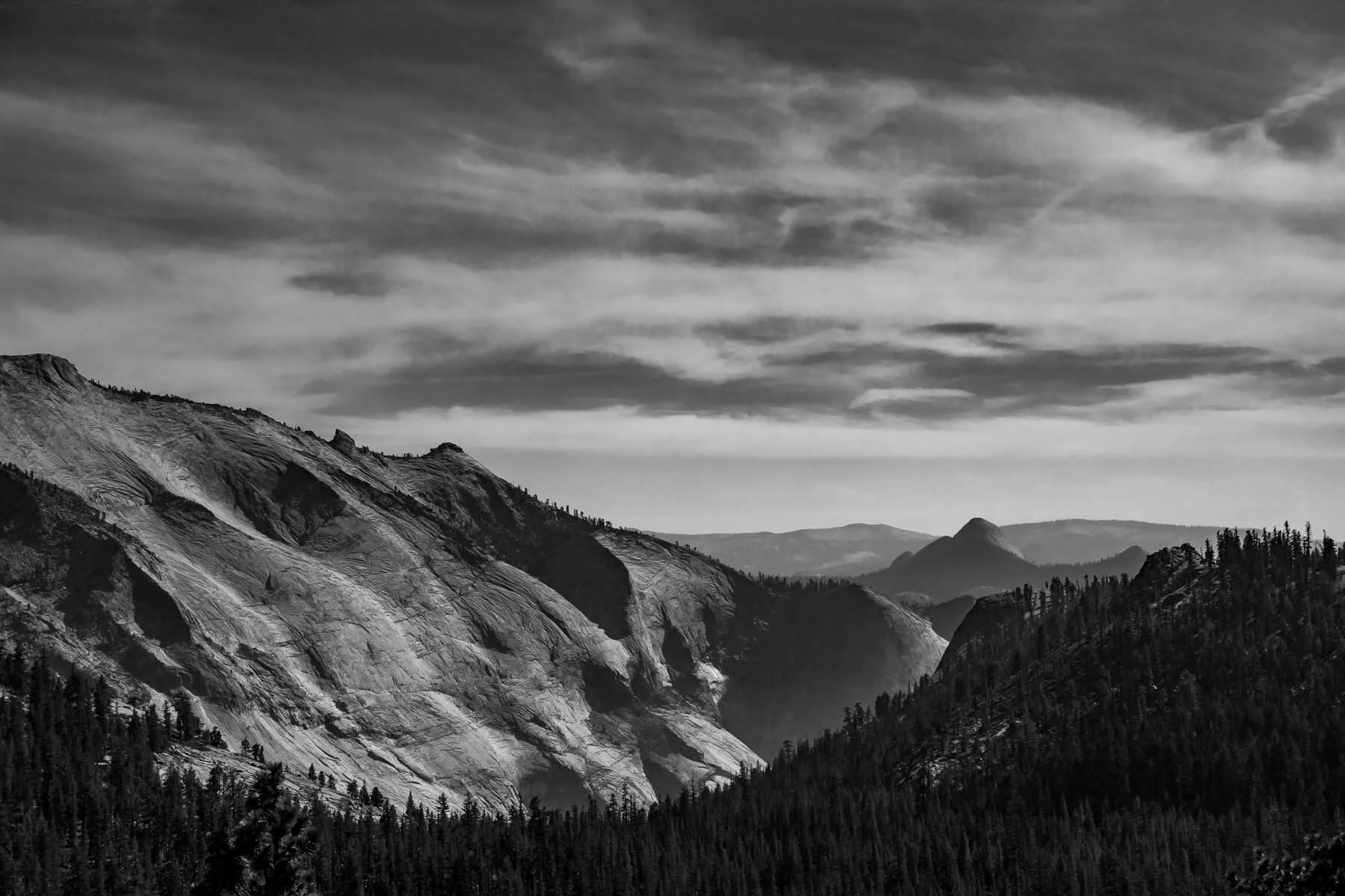 Black and white landscape of mountain range with rocky peaks, forested slopes, and cloudy sky.
