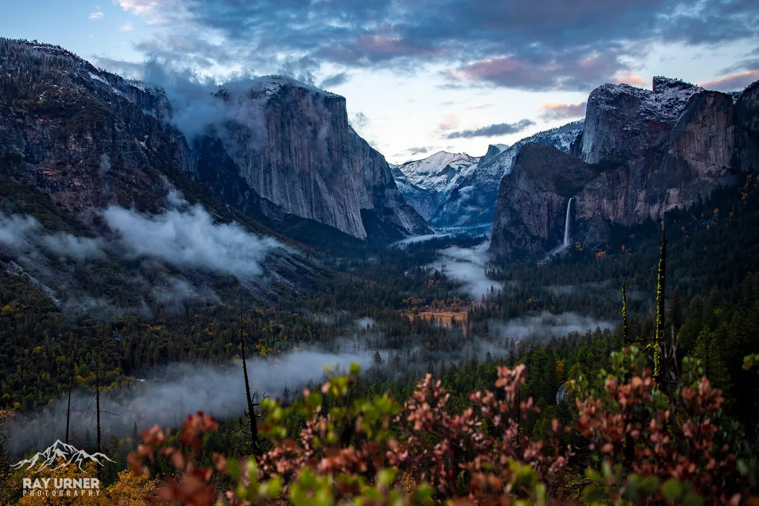 A scenic view of a misty valley with tall mountains, including a waterfall on the right, dense forest, and cloudy sky at dusk.