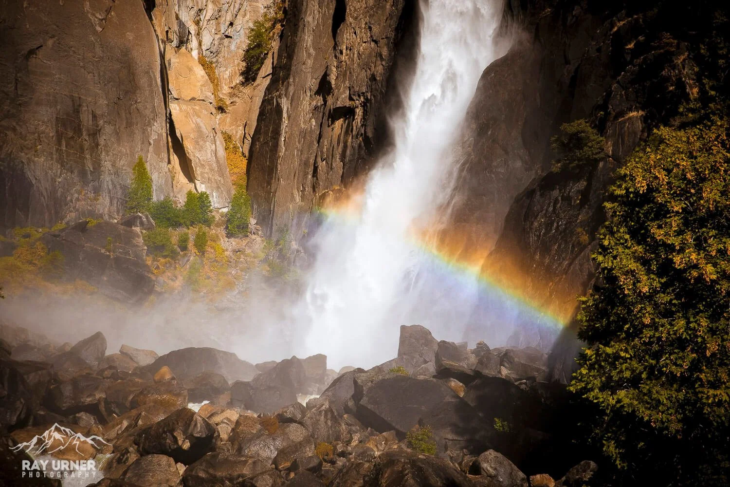 A cascading waterfall flowing between large rugged mountains with mist at the base and a rainbow in the spray.