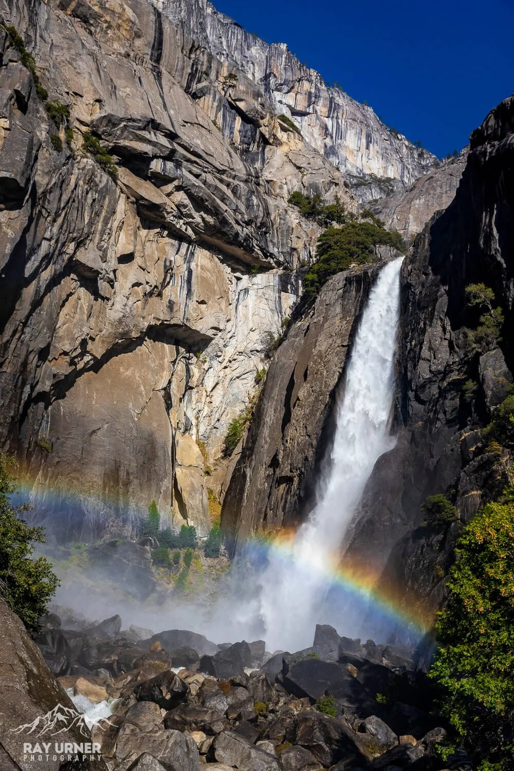 Tall waterfall cascading down rocky cliffs with a rainbow near the base, surrounded by green trees and a clear blue sky.