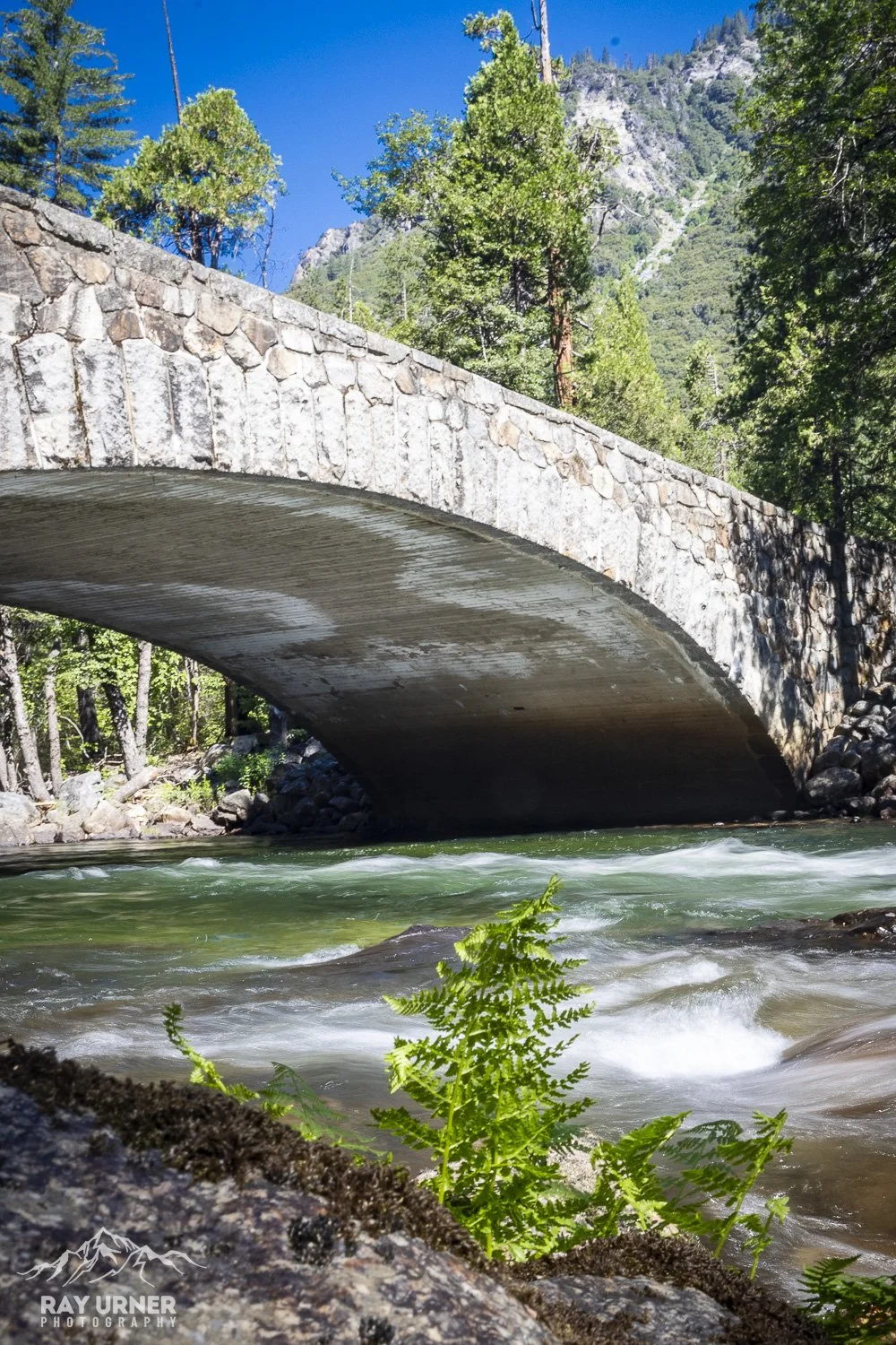 A stone arch bridge over a fast-moving river surrounded by green trees and mountainous landscape under a clear blue sky.