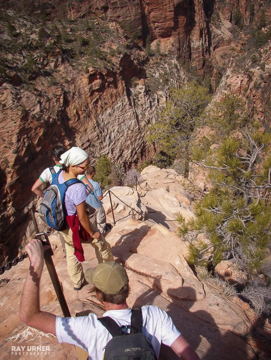 A group of hikers walking along a rocky trail with chains, descending into a canyon with red rock walls and sparse desert vegetation.