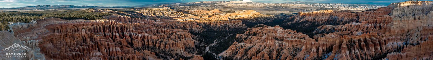 Panoramic view of colorful layered rock formations in a desert landscape, with distant mountain ranges and a partly cloudy sky.