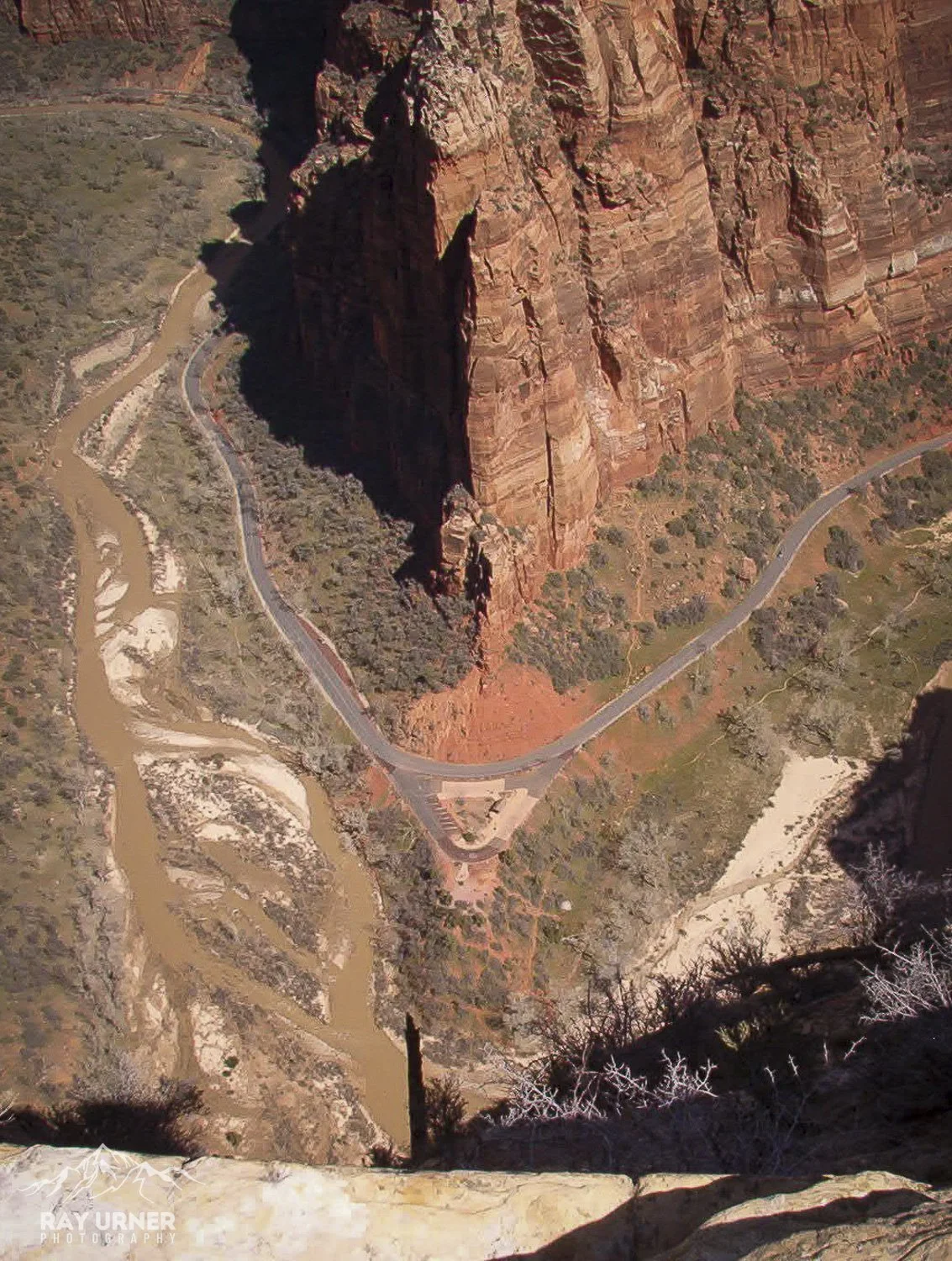 Aerial view of a winding road and a river below a tall red rock canyon, with shadows cast from the canyon walls.
