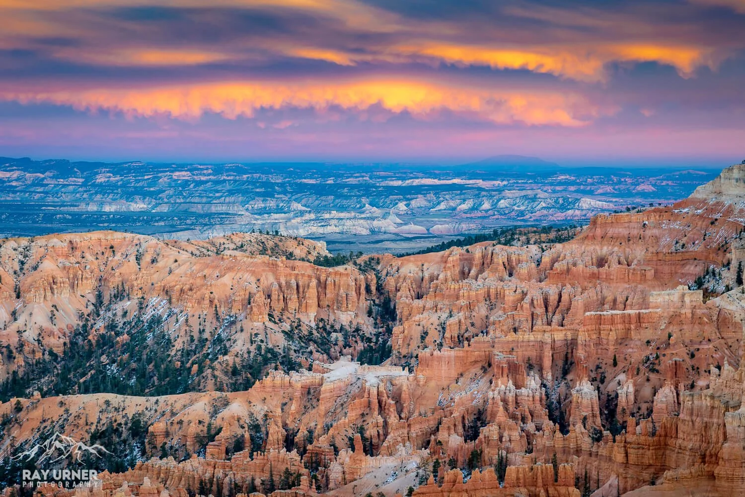 A panoramic view of the Grand Canyon at sunset, showing layered rock formations in orange, pink, and red hues, with patches of snow on the ridges and a colorful sky with clouds.