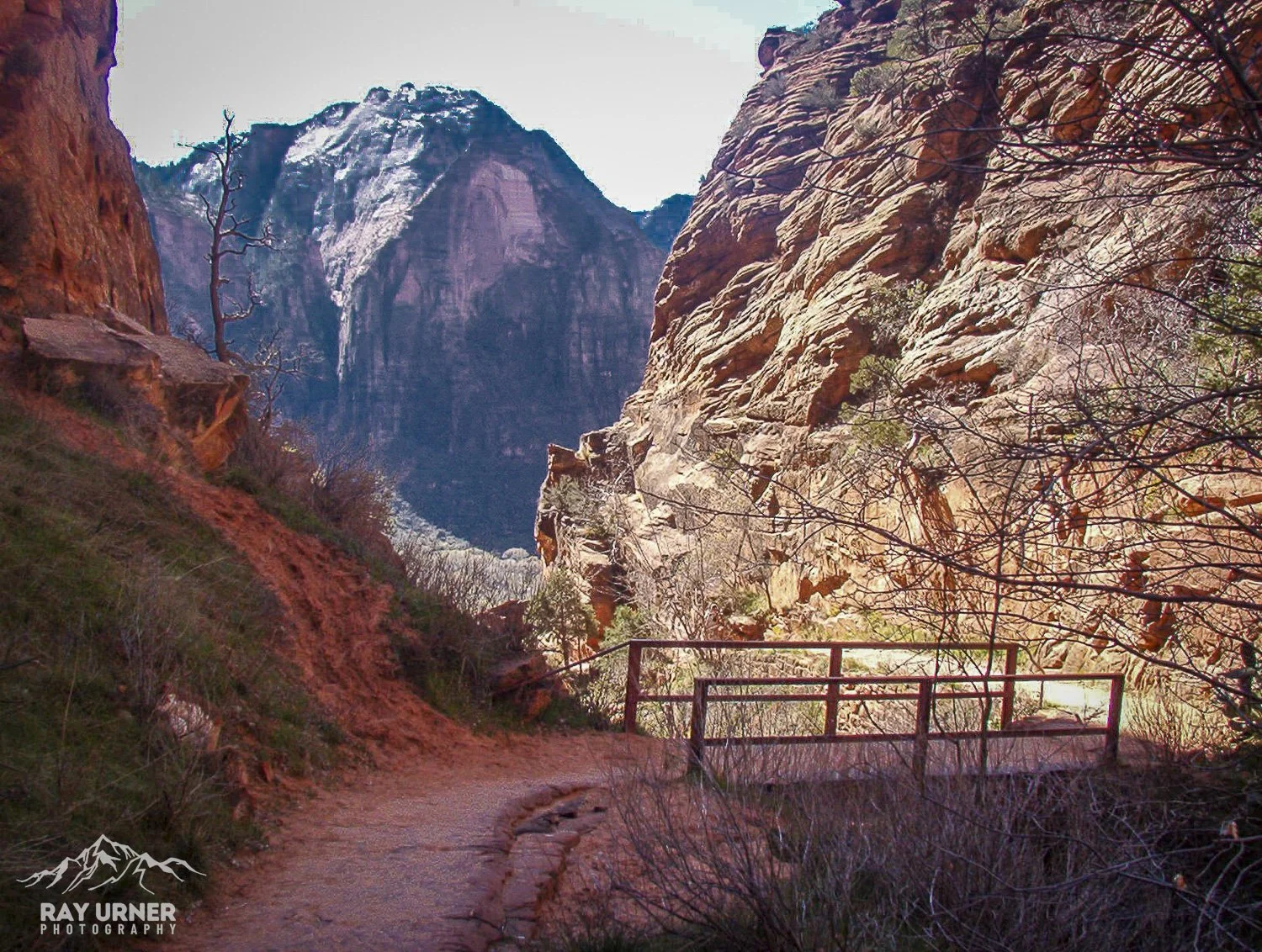 A trail with a small bridge leading into a canyon with large rock formations and a waterfall in the background.