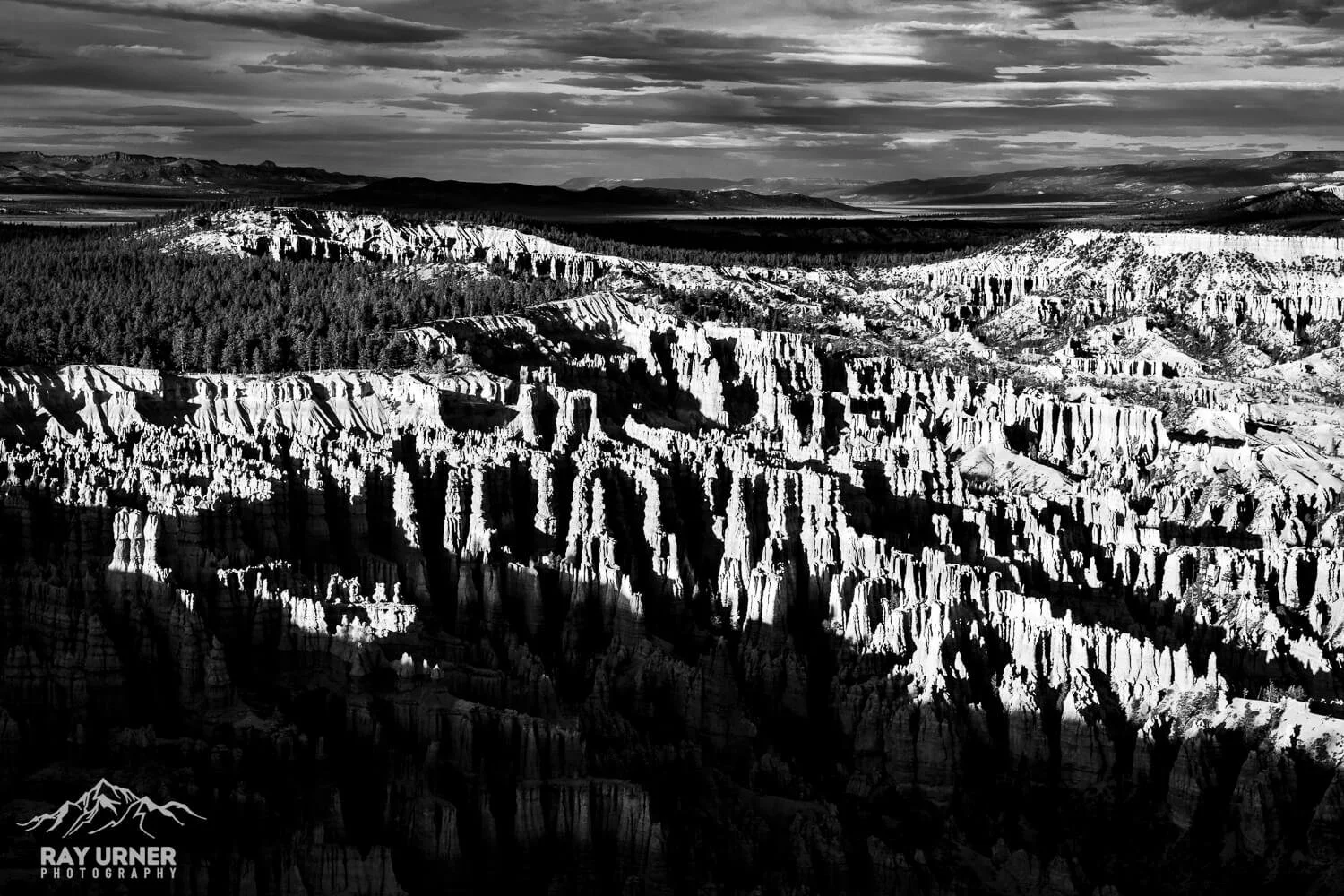 Black and white photograph of a rugged mountain landscape with tall, jagged rock formations and dense forested areas in the background, under a cloudy sky.