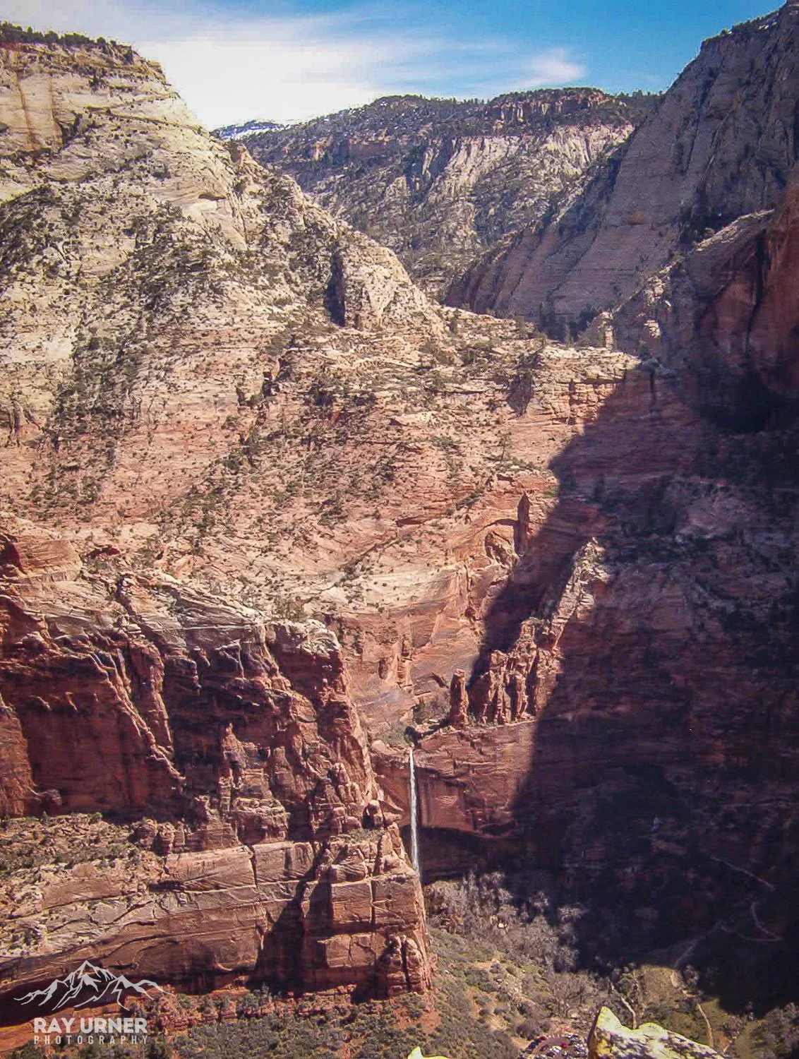 Scenic view of Zion Canyon with tall red and white layered rock cliffs, a small waterfall in the distance, and a partly cloudy blue sky.