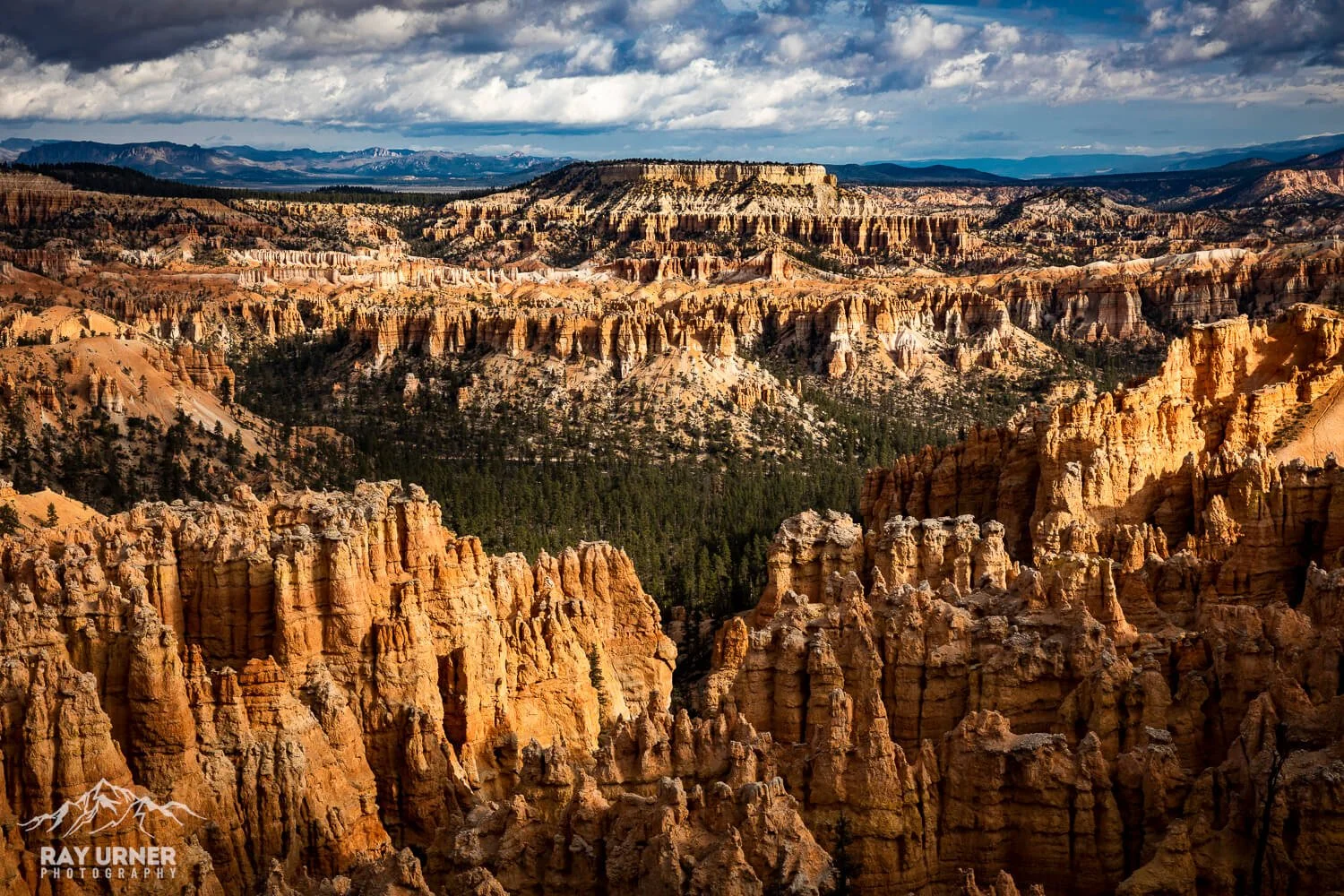 A panoramic view of Bryce Canyon National Park showing colorful hoodoos, layered cliffs, and distant mountains under a cloudy sky.