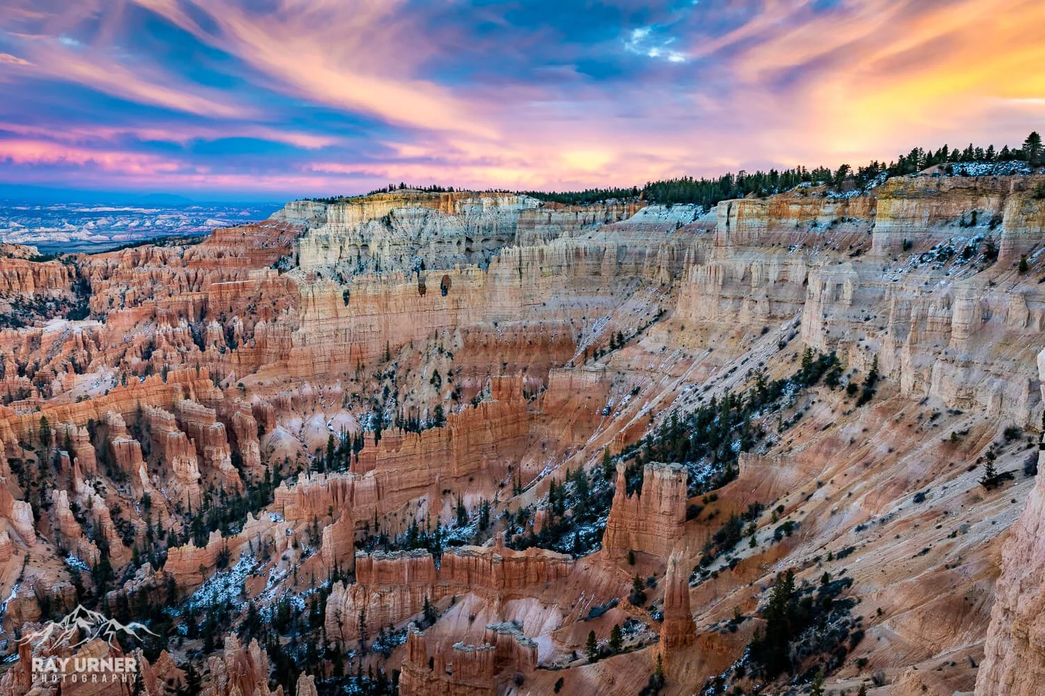 View of the Grand Canyon at sunset with colorful sky, layered canyon walls, some trees, and snow patches.