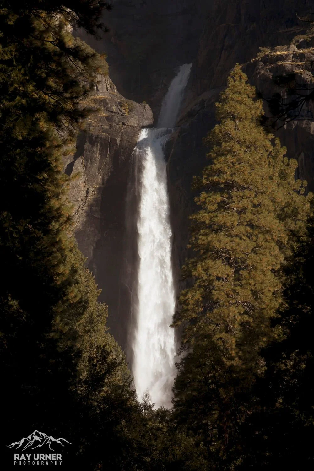 Tall waterfall cascading down rocky cliff into a misty pool, surrounded by green pine trees.