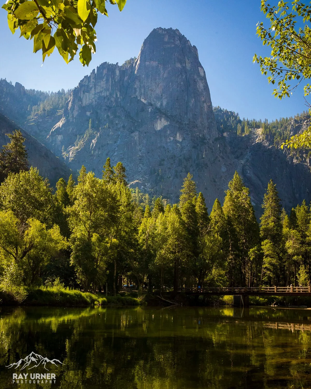 A scenic view of a river reflecting green trees, with a towering granite mountain in the background under a clear blue sky.