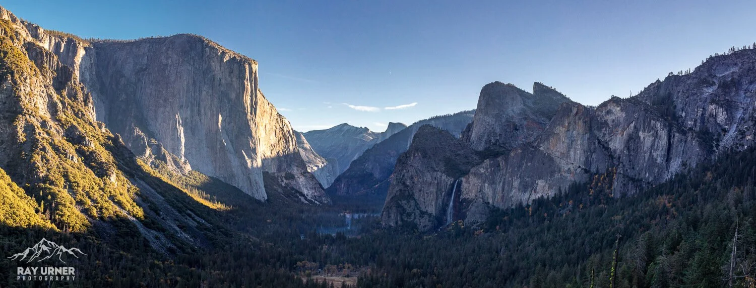 Scenic view of a mountainous canyon with cliffs, dense forest, and a waterfall in the distance, under a clear blue sky.