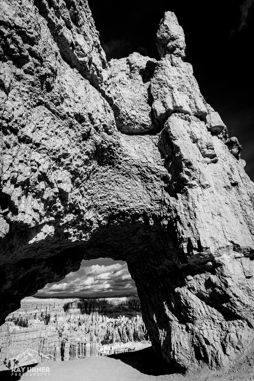A large, rugged rock formation with a natural arch at its base, overlooking a landscape with hoodoos and cloudy sky in the background.