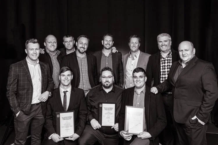 Eleven men in formal attire, some holding framed certificates, posing together in front of a dark curtain at an awards or recognition event.