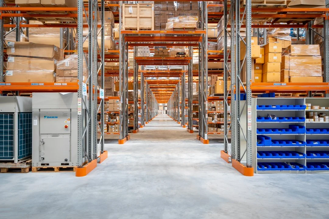 A large warehouse with tall metal shelving units filled with cardboard boxes and blue plastic bins, organized in an aisle.