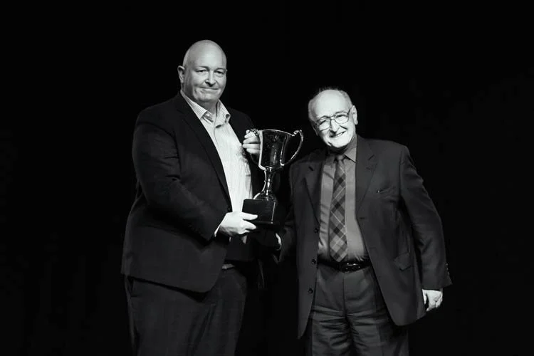 Two men in suits holding a trophy together, standing on a dark stage.