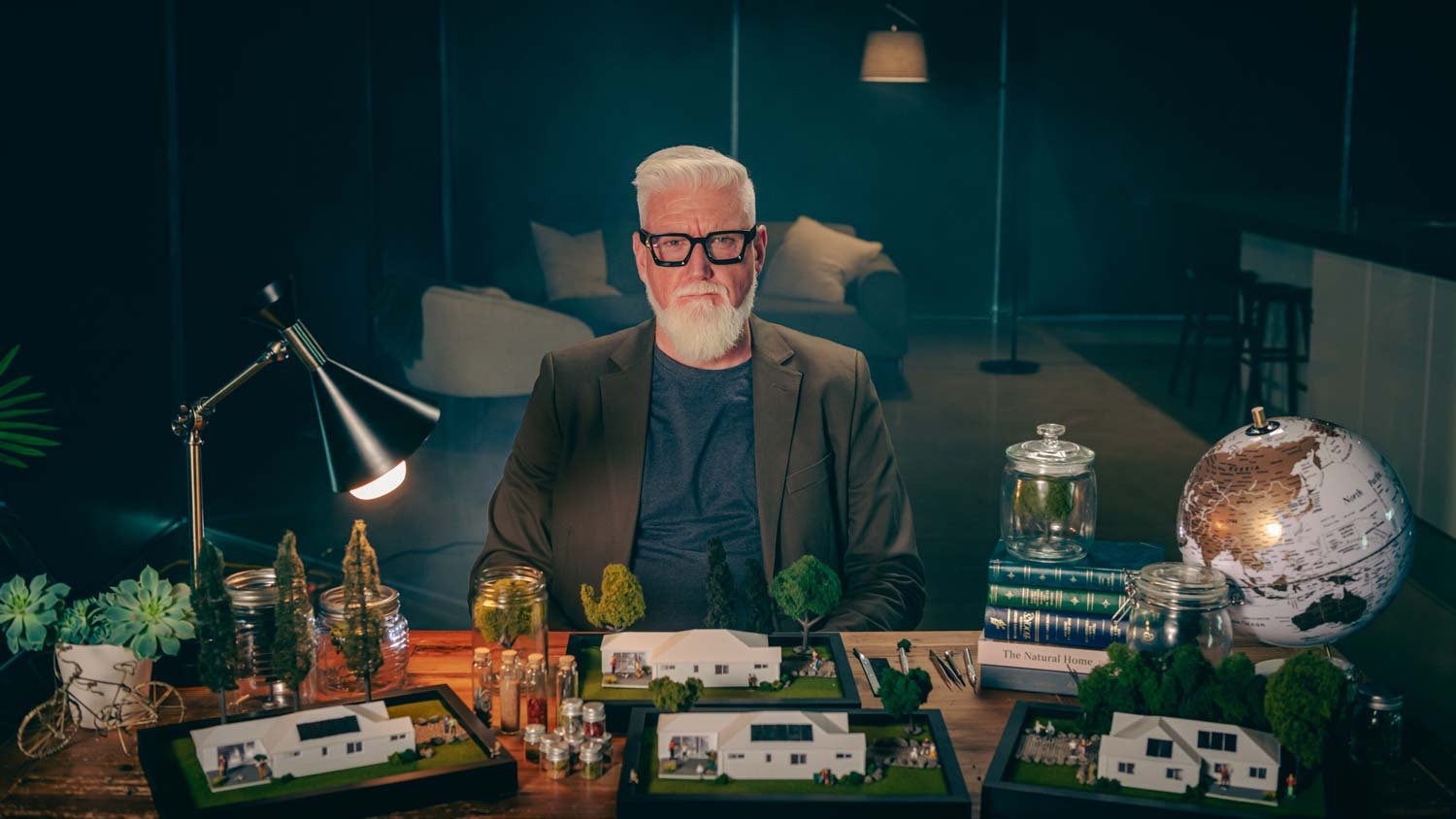 An elderly man with white hair and beard, wearing glasses, a dark blazer, and a blue shirt, seated behind a desk with architectural models, jars, and books. A globe, a lamp, and plants are also on the desk, with a glass wall and a living room visible