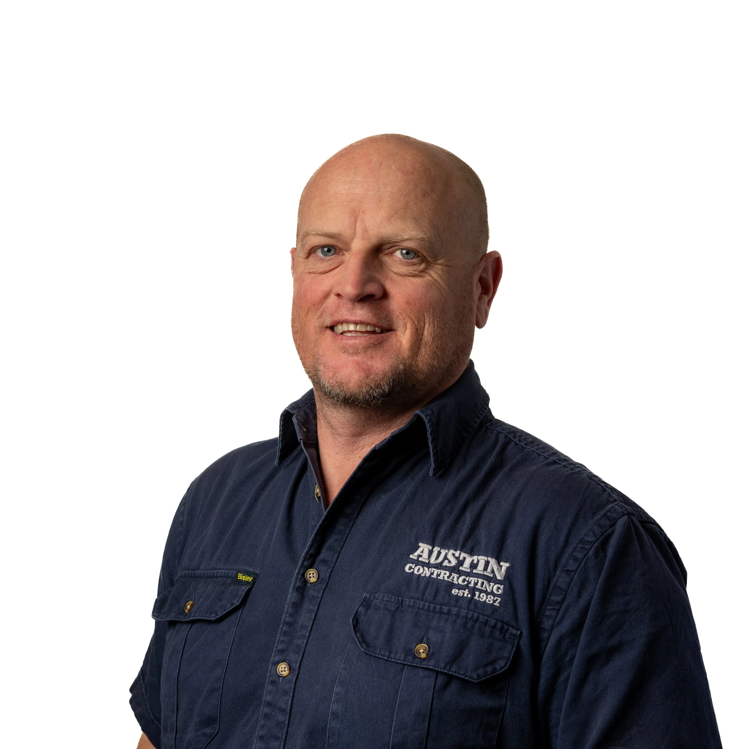 Close-up of a bald man with blue eyes wearing a dark blue shirt with embroidered text on the left chest area, smiling slightly, against a white background.