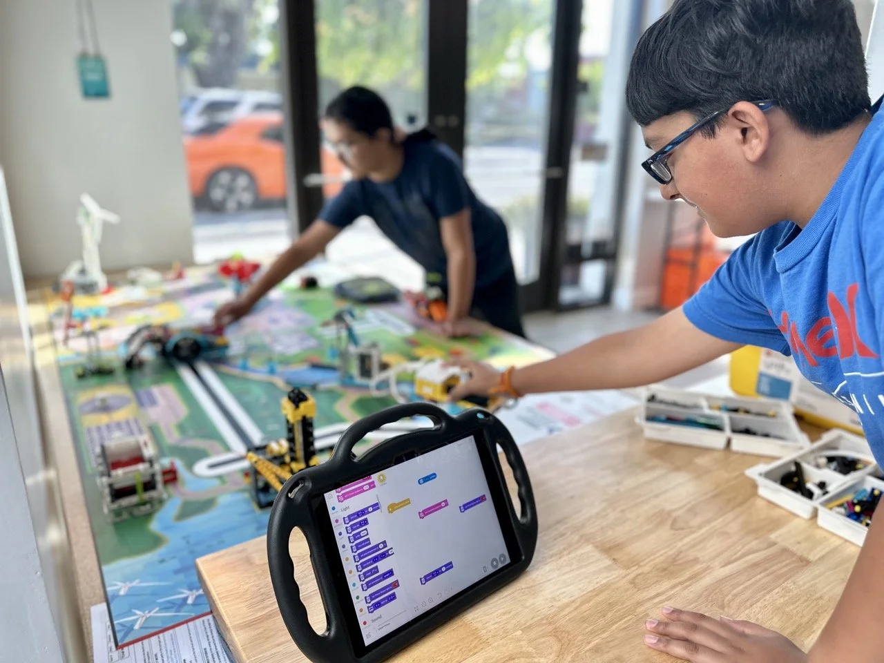 Two students collaborating in a FIRST LEGO League challenge; one is coding on an iPad while the other arranges mission models on the competition field.