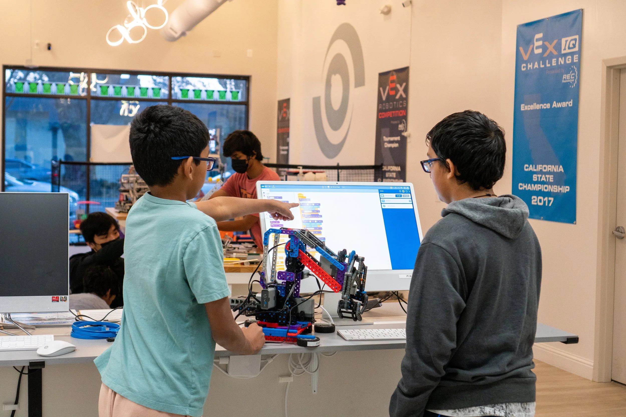 Two students collaborating on VEX IQ robotics programming, with one pointing at code on a laptop screen while their crane robot sits between them on the table.