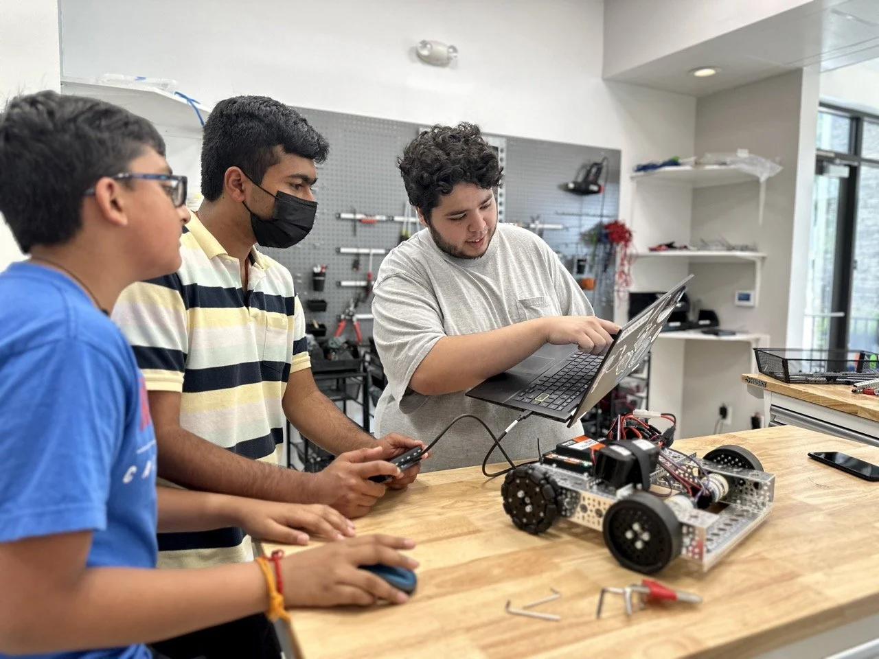 A robotics coach discussing the programming of a FIRST Tech Challenge robot with two engaged students, reviewing autonomous and teleoperated functions.