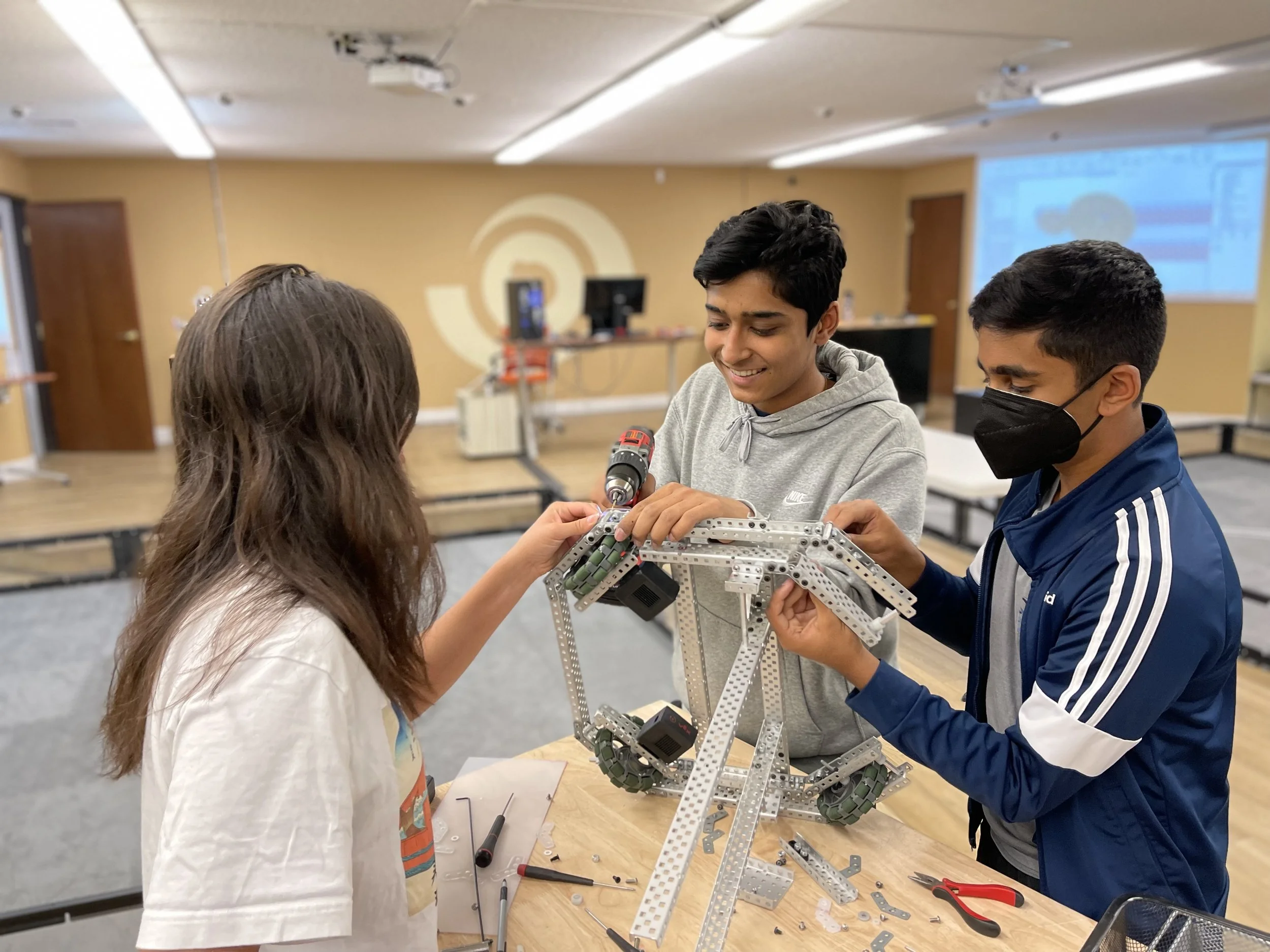 Three students enjoying a hands-on robotics experience, working together on their VEX robot and smiling as they problem-solve.