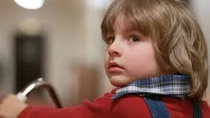 Danny Torrance pauses in a quiet hallway, sensing something unseen, which highlights his psychic ability and the trauma behind his character.