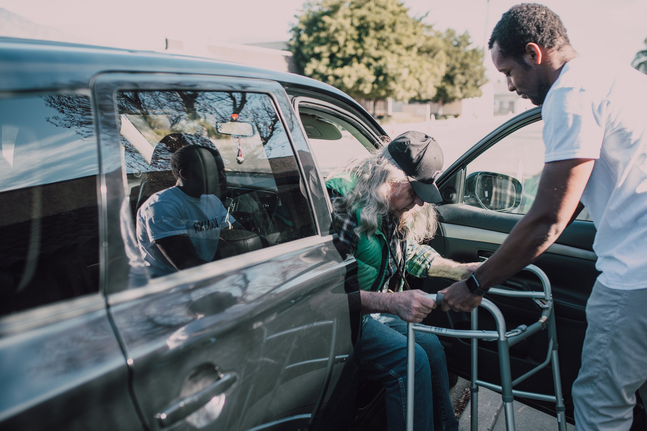 A young man helping an elderly woman with mobility issues in the car while she is using a walker.