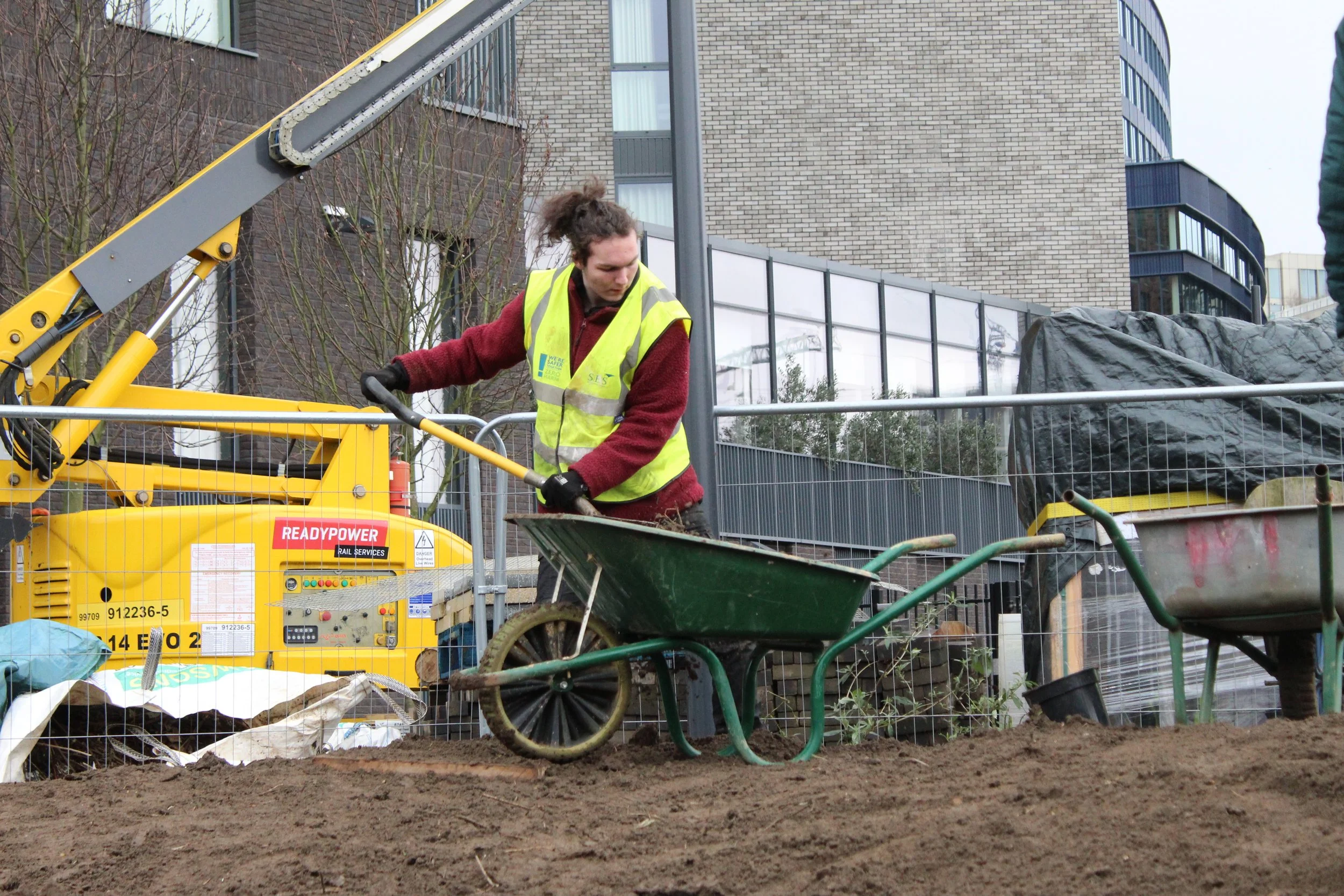 a young person in a high visibility vest uses a spade to move soil from a wheelbarrow unto the ground