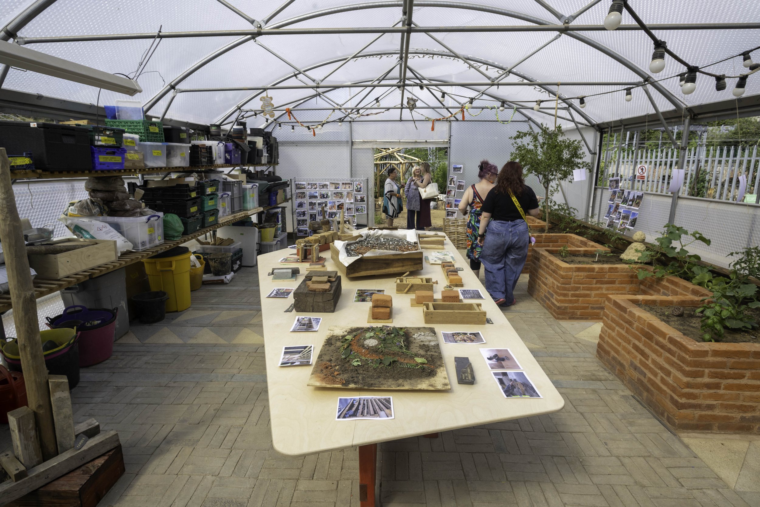 The interior of a paved polytunnel with brick planters to the right and shelving to the left, a table sits in the middle of the space