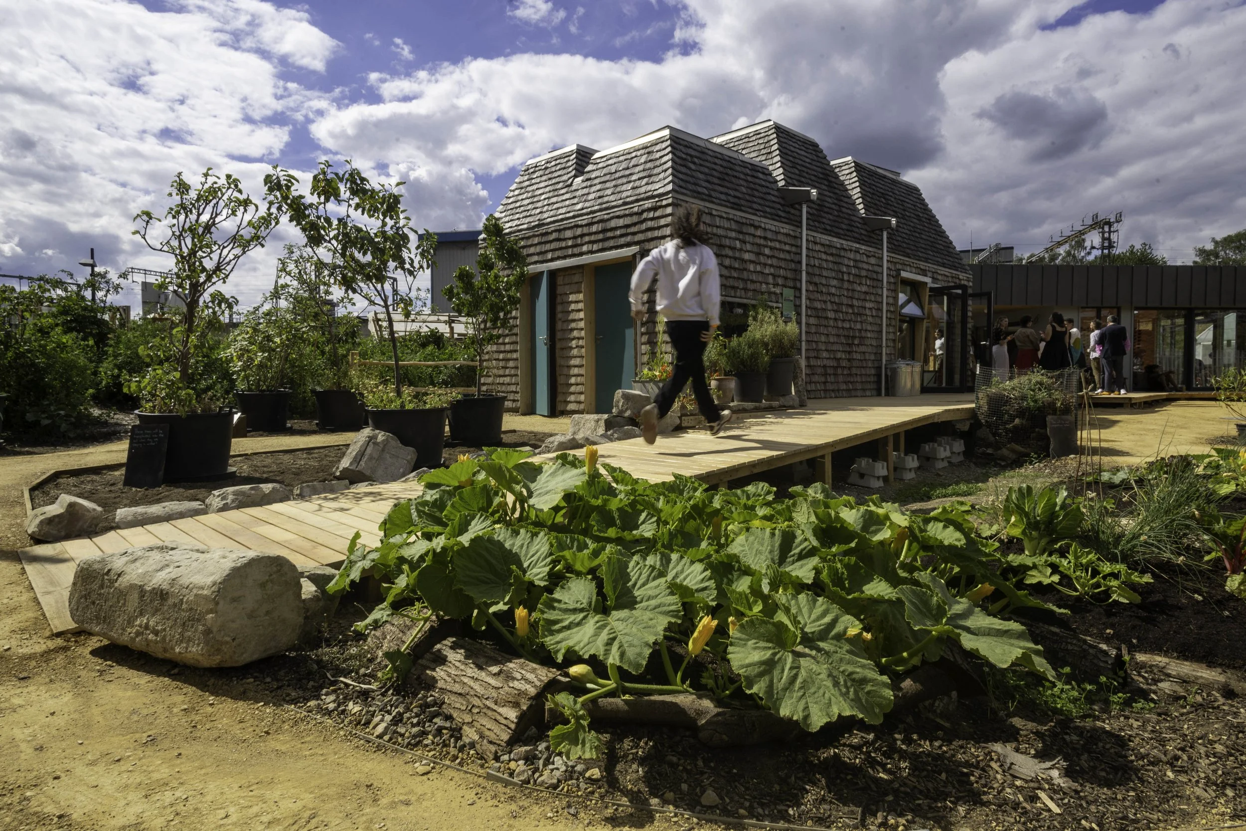 A child runs up a ramp towards a building. There are plants and trees around the ramp and the sun shines brightly