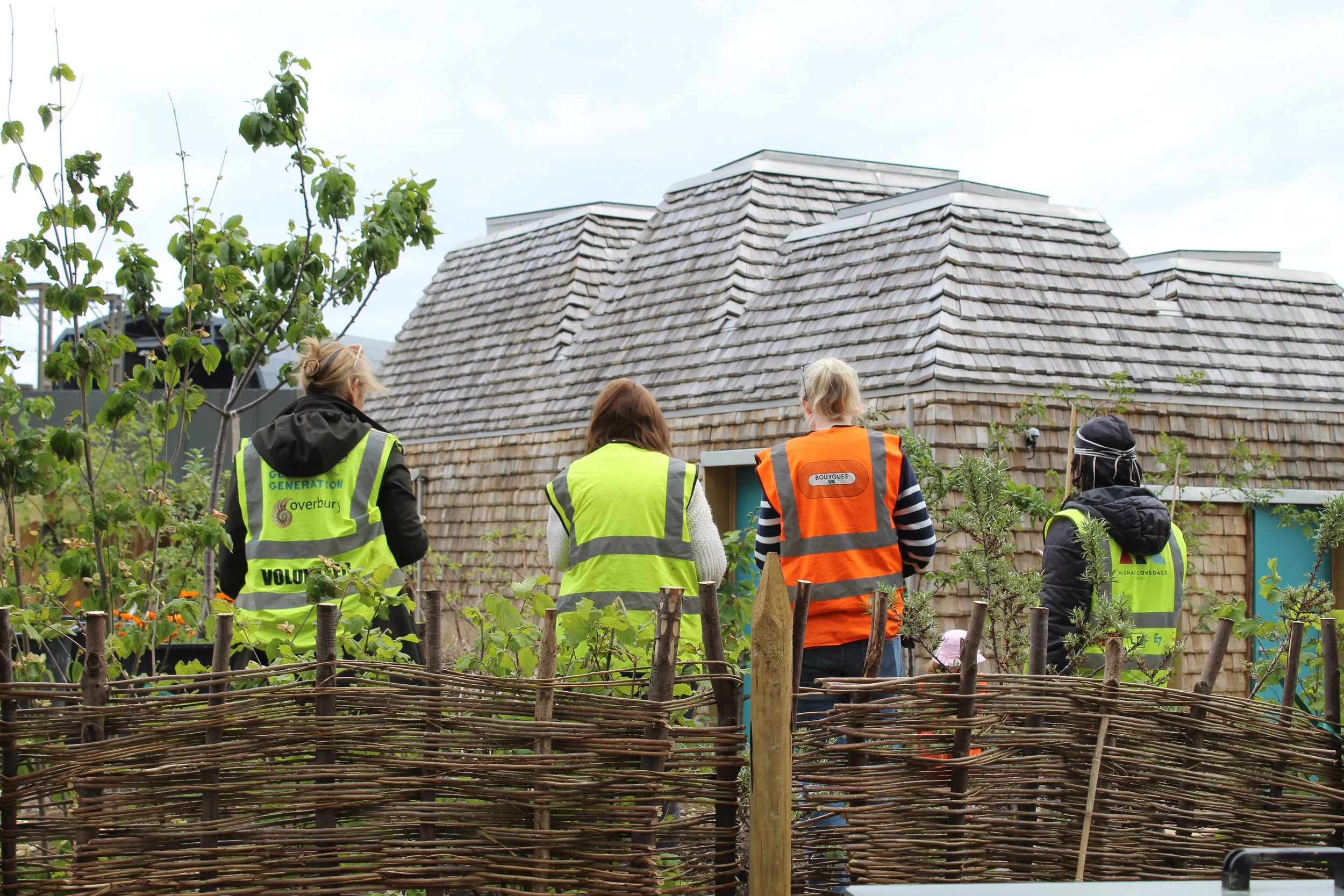 Four people in high visibility vests stand with their backs to a willow fence and face a building covered in chestnut shakes.
