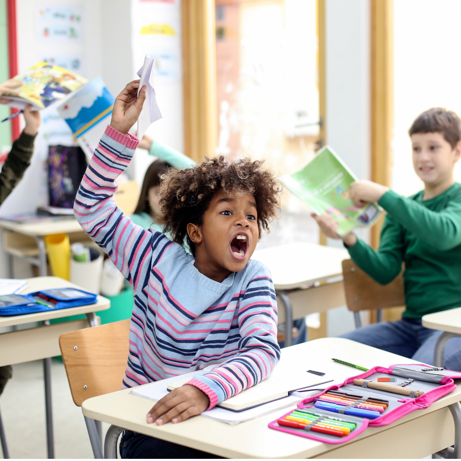 A diverse group of children in a classroom, with a girl in the foreground raising her hand and shouting, while other children read and work at their desks.