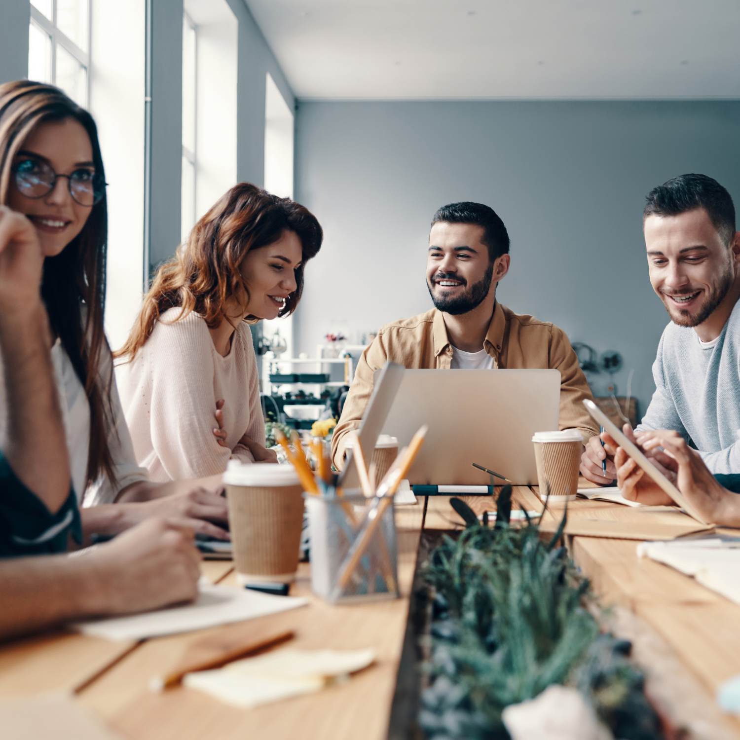 A diverse group of five young professionals sitting around a wooden table in a modern office space, engaged in a meeting or collaborative work, with laptops, tablets, notebooks, and coffee cups on the table, and a large window in the background letting in natural light.