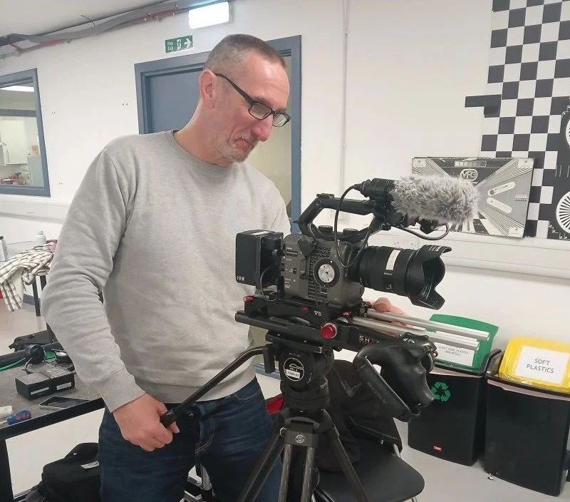 An older man with glasses, dressed in a grey sweatshirt and blue jeans, operates a broadcasting camera in a training room.